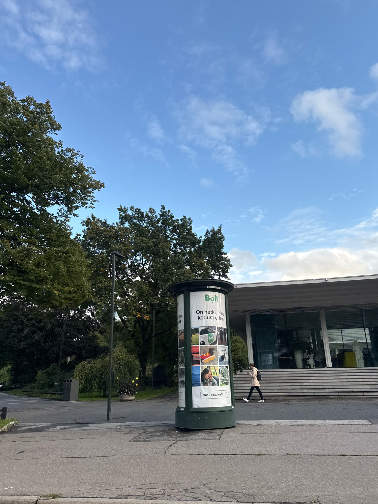 The image shows a bright, cloudy sky over a street scene in Tallinn, Estonia. In the foreground, a cylindrical advertising pillar displays various images, including a Bolt logo and a car. To the right, a modern building with large windows and a tiered entrance is visible. A person wearing a light-colored jacket and dark pants walks across the street in front of the building. Green trees and a lamppost are situated to the left of the advertising pillar, with a park-like area in the background containing a trash can and a bench.
