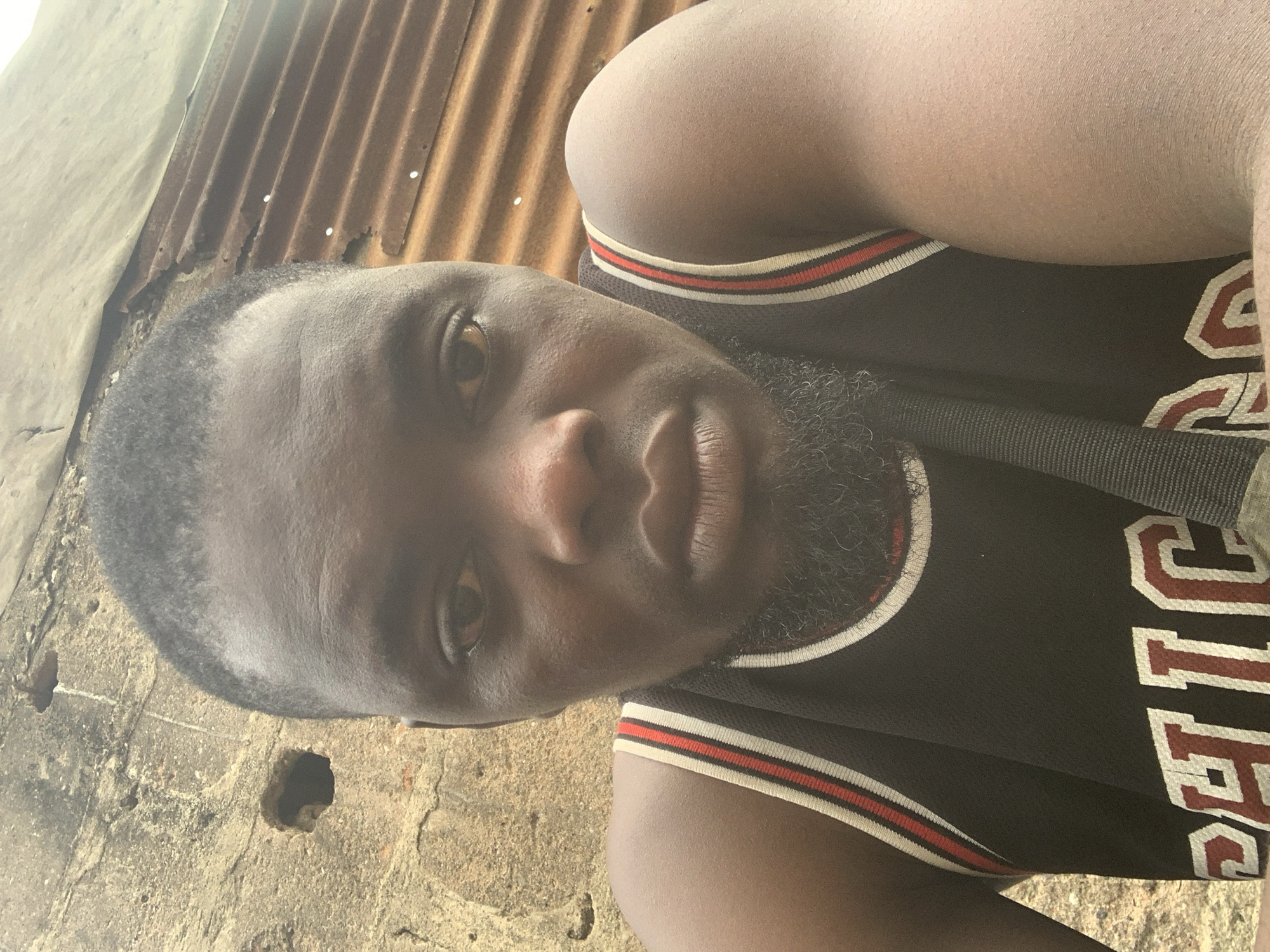 The image is a selfie taken outdoors, likely in a weathered urban or semi-urban environment. The subject is a Black male, appearing to be in his late twenties or early thirties, with dark skin, a closely cropped hairstyle, and a beard. He is wearing a black basketball jersey with red and white trim. The jersey has white and red lettering that appears to read "CHICAGO" on the front.
The background is divided. To the left is a rough, textured wall made of concrete or stucco, showing signs of age and wear, with a visible hole in the upper left portion. To the right is a corrugated metal surface, appearing rusty and weathered, possibly part of a roof or a fence. The lighting suggests it is daytime, with a bright light source, possibly the sun, casting some glare.
While the image does not contain explicit markers of Shagamu, Nigeria, the overall aesthetic of the weathered concrete and corrugated metal structures is commonly found in many parts of Nigeria, including Shagamu. The image captures a casual, unposed moment of the subject.