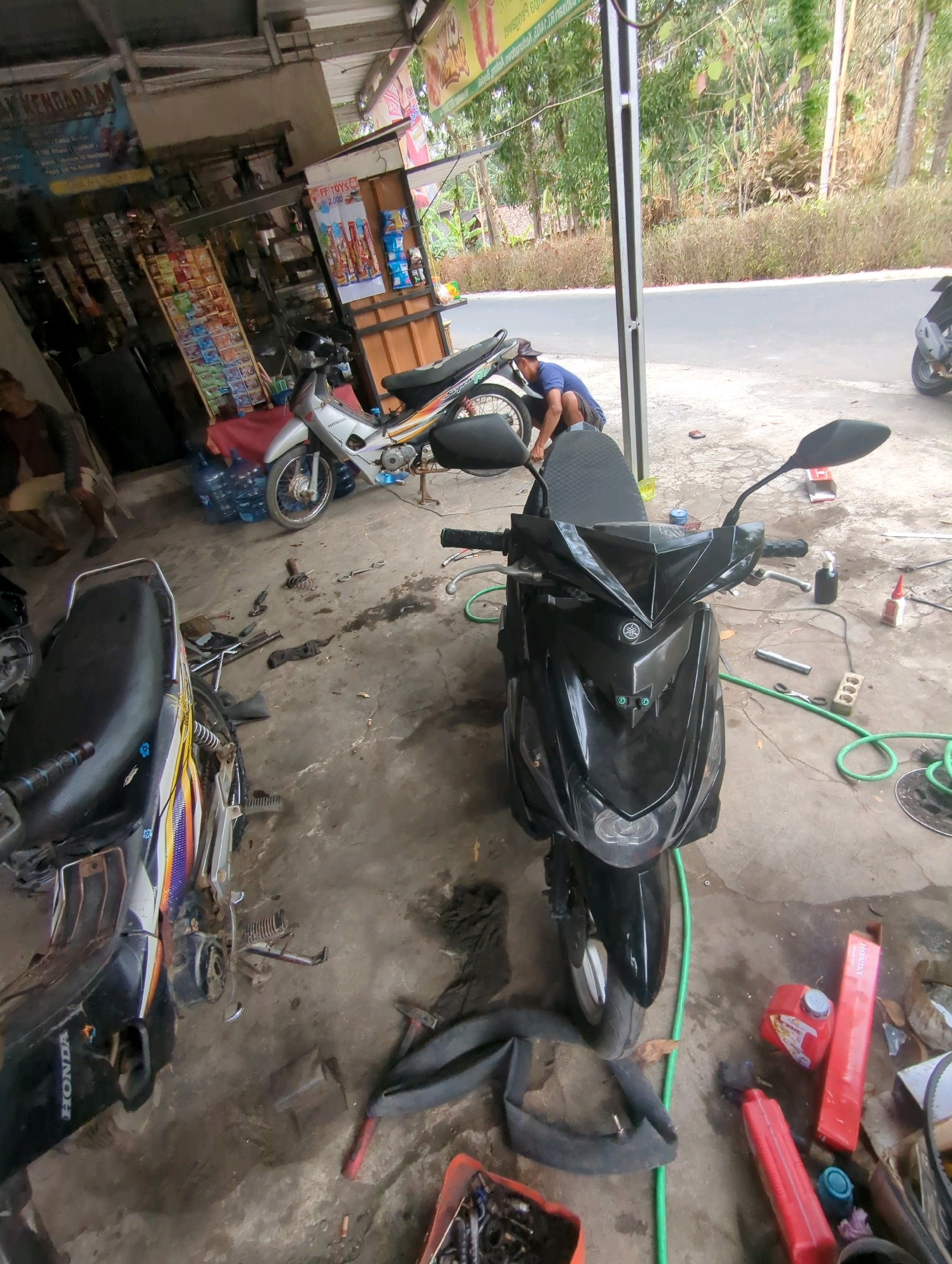The image depicts an outdoor motorcycle repair shop, likely in Kedungdowo, Indonesia. Two motorcycles are present: a silver and orange motorcycle is parked in the background, and a black scooter is positioned in the foreground. A person, wearing a blue t-shirt and dark shorts, is crouched down and appears to be working on the rear wheel of the silver motorcycle. Various tools, including a wrench, a spring, and a black cloth, are scattered on the concrete ground around the motorcycles. In the background, shelves are stocked with goods, and a road is visible with trees and vegetation lining it. The scene suggests a typical day at a local mechanic's workshop.