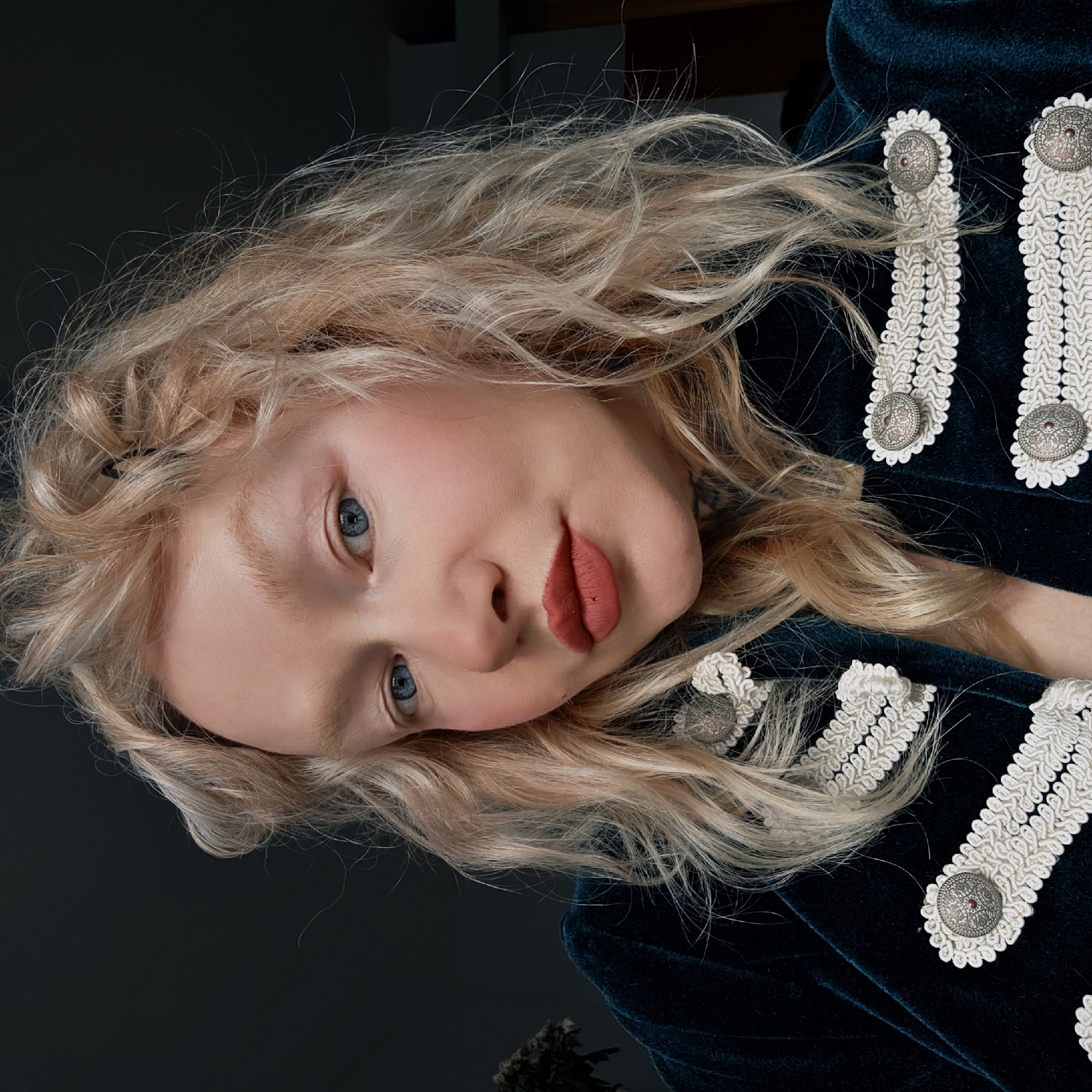 The image shows a fair-skinned person with blonde, wavy hair and striking blue eyes, tilted so their face is near the center of the frame. They have high cheekbones and are wearing matte red lipstick. They are wearing a dark teal velvet jacket adorned with decorative white trim and ornate silver buttons. The background is a dark, muted gray color. Based on the style of the jacket and overall aesthetic, there is no indication of the location being Saint Petersburg, Russia.