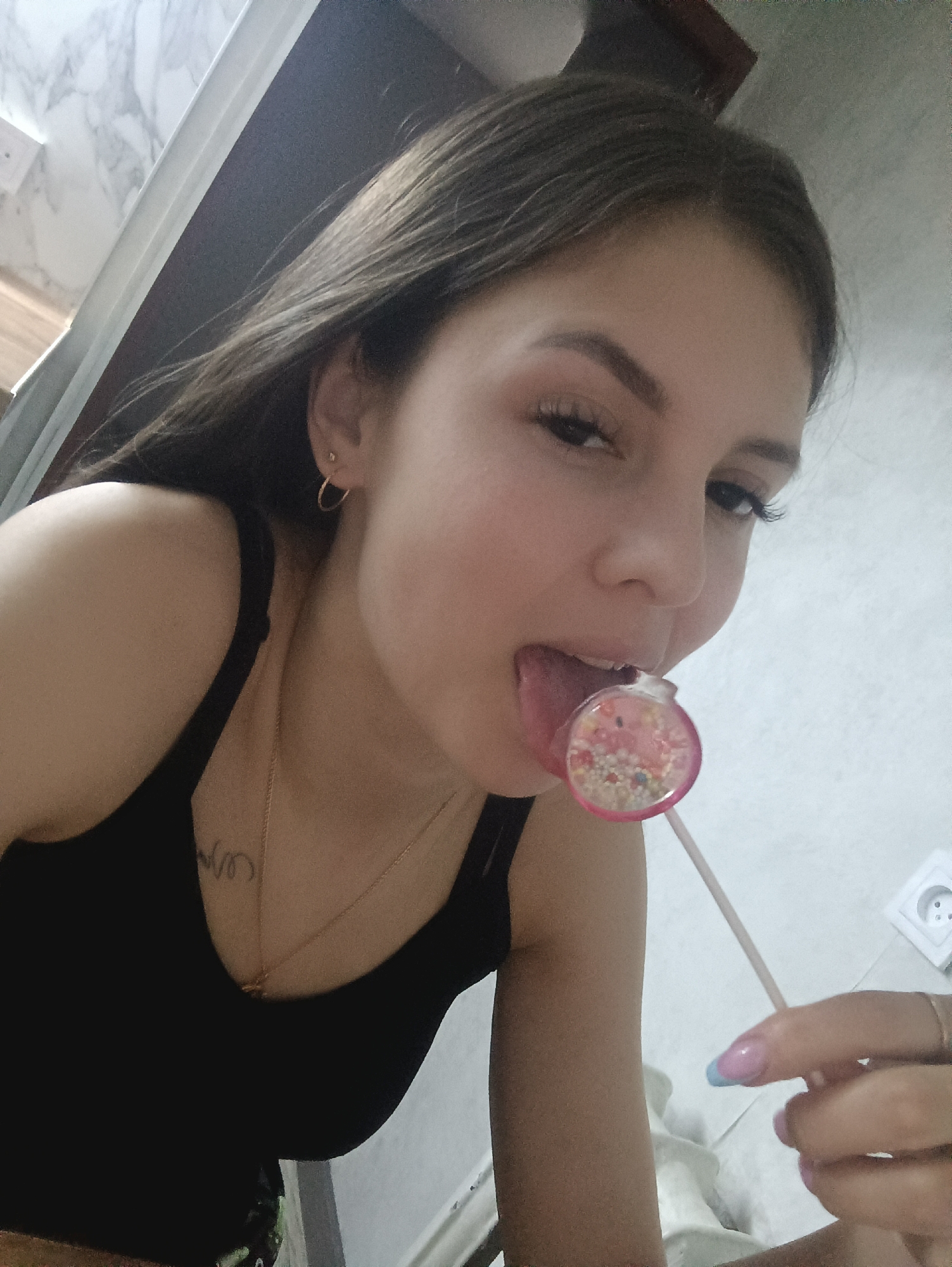 A young woman with long brown hair, fair skin, and manicured nails (pink and blue) is indoors, holding a pink lollipop to her mouth. She is wearing a black tank top and a gold necklace. There is an electrical outlet on the wall behind her, and what appears to be marble-patterned tiling or wallpaper is visible in the upper-left corner. A tattoo is visible on her upper chest. The location is Unknown, Unknown.