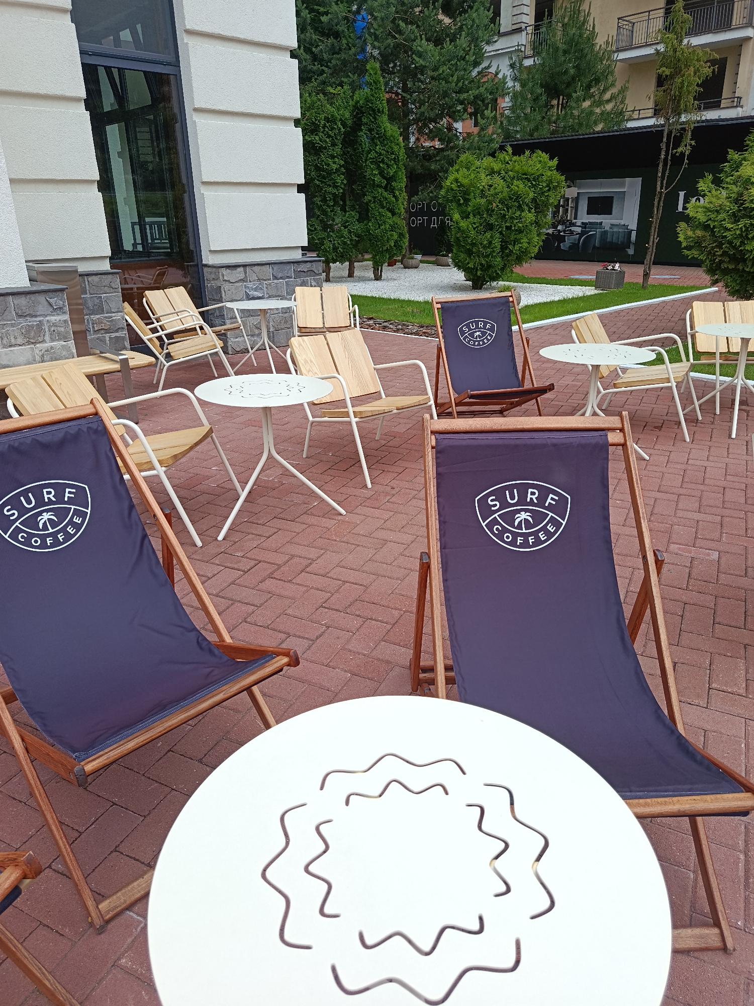 This is a street-level, eye-level, medium shot photograph of an outdoor seating area of a cafe. The scene is set in Estosadok, Russia. The foreground features a white round table with a decorative wavy pattern cut out of its surface. To the left and right of this table are two dark blue deck chairs with the "SURF COFFEE" logo printed on them in white. These chairs have wooden frames.
In the midground, more wooden chairs and white metal tables are visible. The tables are round and have slender, tri-legged bases. The chairs appear to be made of light-colored wood with metal frames. The ground is paved with red brick pavers arranged in a herringbone pattern.
The background shows a modern building with large glass windows and a facade constructed of textured light-colored blocks and a stone base. Beyond the building, there are several tall, slender evergreen trees and some bushes. In the far background, a multi-story building with balconies is visible, suggesting an urban or resort setting. A large advertisement banner with an image of a living room is also present, partially obscured by trees. The overall impression is that of a cafe or restaurant outdoor dining space, likely in a resort area given the lush greenery and architectural style. There are no people visible in the image.