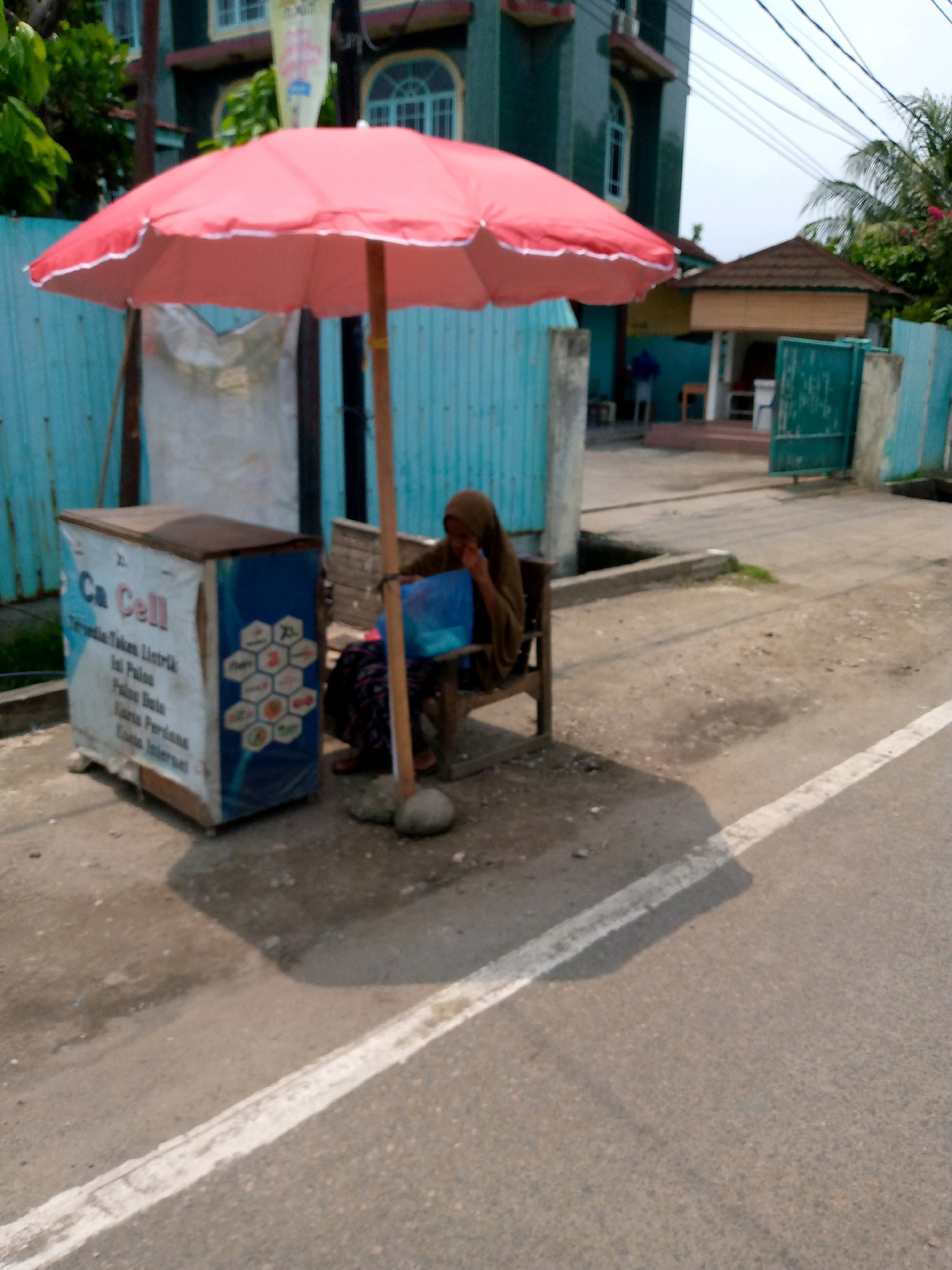 The image depicts a street scene in Lhokseumawe, Indonesia, possibly near a residential or commercial area with a building in the background. In the foreground, a red umbrella shades a small kiosk, identified by the text "Ca Cell" on its side. A woman, wearing a hijab and brown clothing, sits on a wooden bench next to the kiosk, appearing to be tending to something in a blue container. The kiosk itself appears to be a mobile stand, possibly for selling phone services or related items, with various graphics and text on its exterior. The street is paved, with a white line marking the edge of the road. In the background, a teal-colored fence and a building with arched windows are visible. Palm trees suggest a tropical climate. The overall scene suggests a casual, everyday street vendor setup.