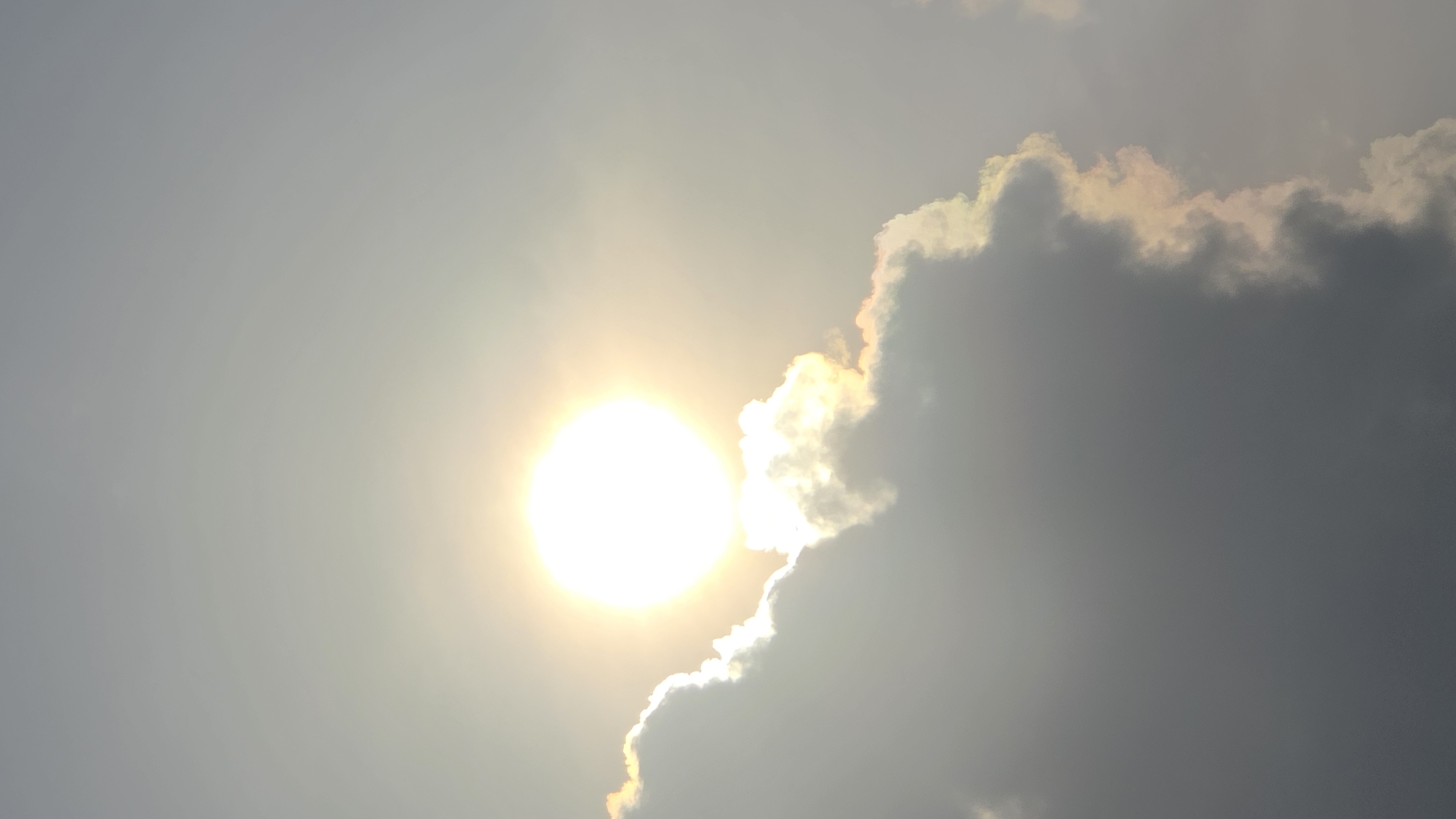 The image depicts the sun obscured by clouds, with bright sunlight radiating through the thinner areas and illuminating the edges of the darker clouds. The background appears to be a hazy sky. No people, specific objects, actions, or interactions are visible. There is no discernible information to confirm the location as Bac Nha Trang Ward, Vietnam.