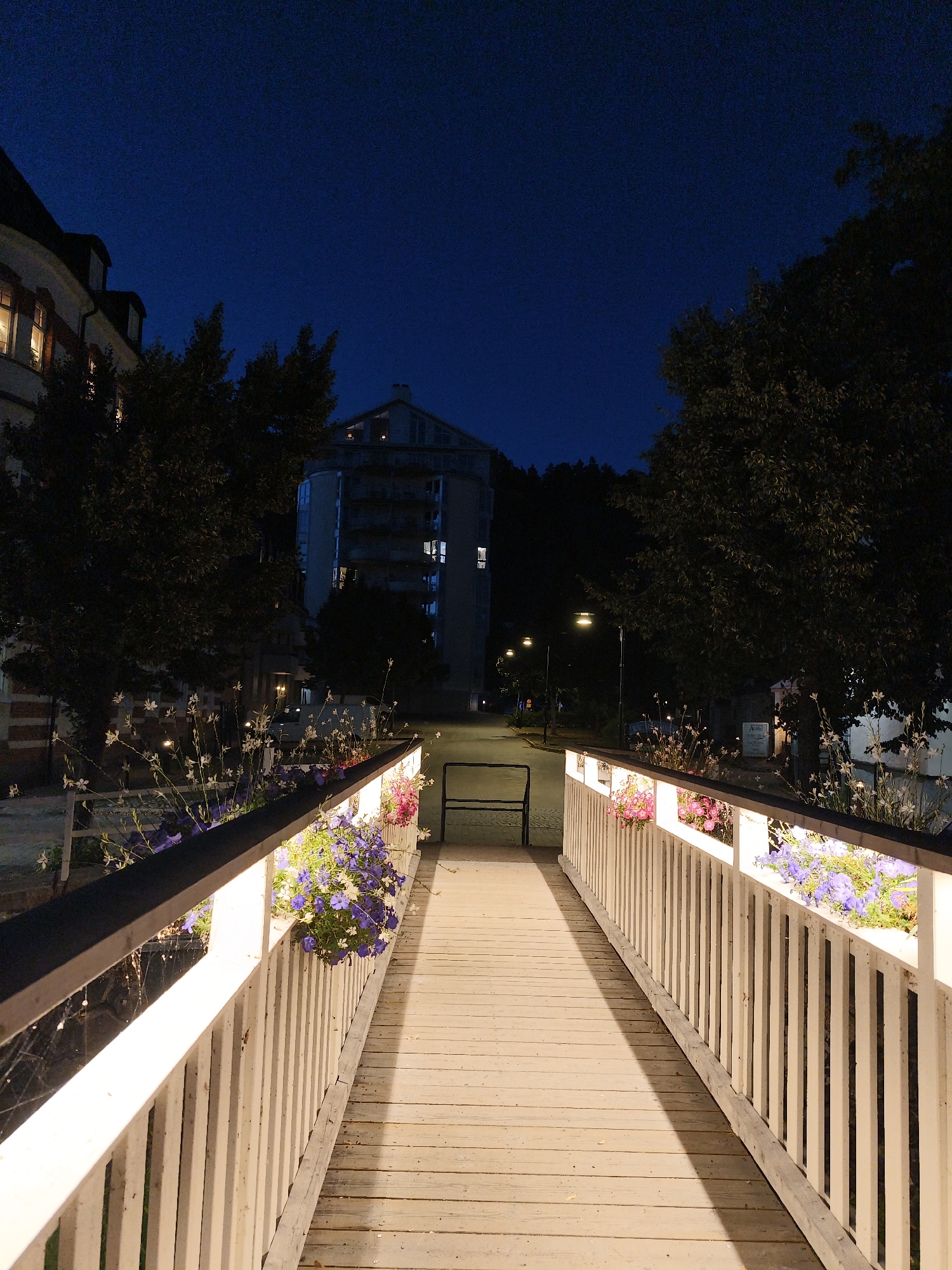 The image depicts a wooden pedestrian bridge illuminated by embedded lights along its railings. Planters filled with purple and pink flowers adorn the railings on both sides of the bridge. The bridge leads towards a building with multiple stories and illuminated windows, and in the background, trees and streetlights are visible under a dark blue night sky. The scene appears to be in the city of Söderköping, Sweden, given the architectural style and the serene nighttime ambiance. There are no people visible in the frame.
