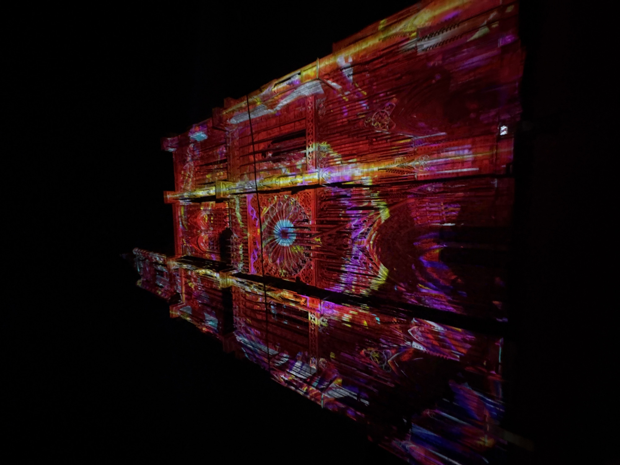 The image shows the Cathédrale Notre-Dame de Strasbourg in Strasbourg, France, illuminated at night with a vibrant projection mapping display. The facade of the Gothic cathedral is covered in a dazzling array of colors, including reds, purples, blues, and yellows, creating intricate patterns and designs. The light show emphasizes the architectural details of the building, such as the arched windows and spires. The sky above is completely dark, highlighting the illuminated cathedral. The perspective is from below, looking up at the towering structure. There are no visible people or cars.