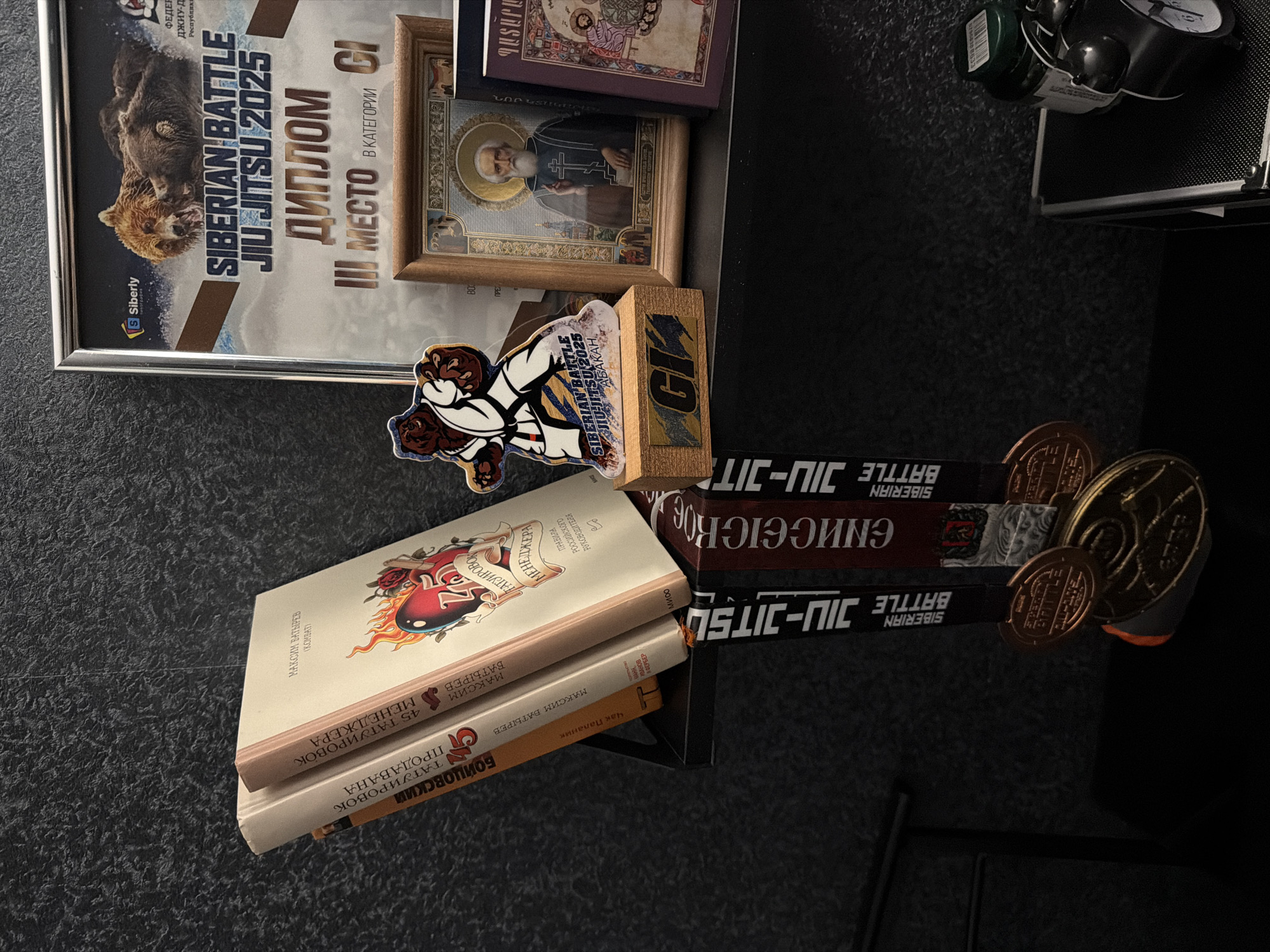 The image shows a collection of items on a dark surface, likely a shelf or table. There is a framed "Siberian Battle Jiu-Jitsu 2025" diploma, signifying third place in a category, located in Abakan. There are two books on top, one titled "Manager's Tattoos" and the second is called "Fighting Club". An icon is also visible, alongside the Jiu-Jitsu belts and medals from the same event, presumably related to the diploma. The presence of both religious and martial arts items suggests a blend of spiritual and combative interests. The location cannot be determined by the image.
