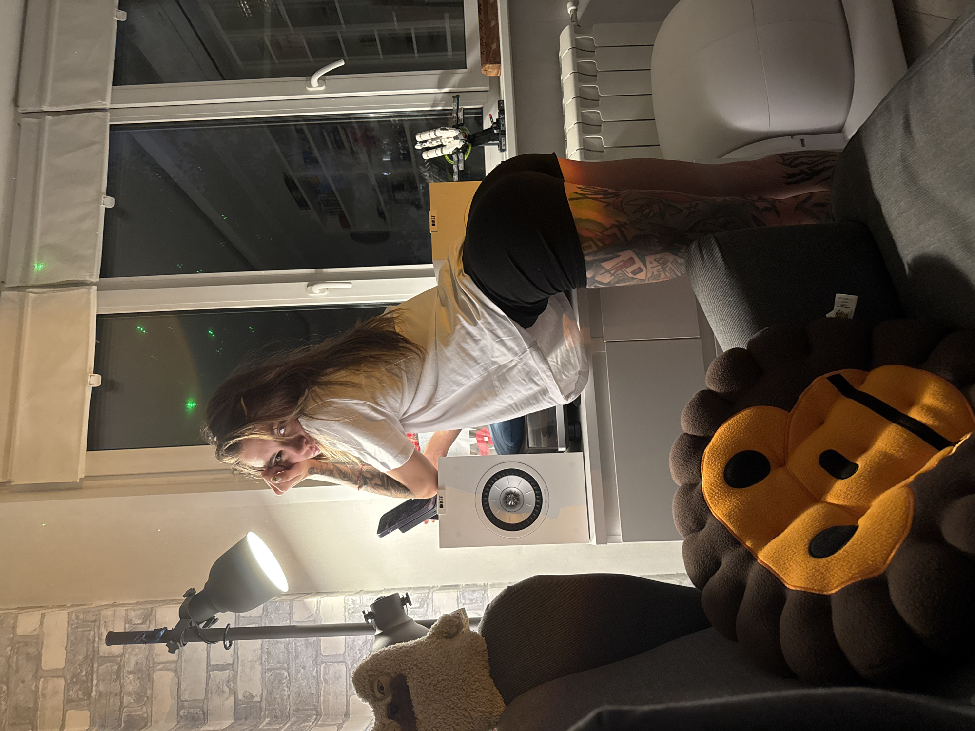 A young woman with extensive tattoos on her arms and legs is leaning over a white cabinet. She is wearing a white t-shirt and black shorts. In the background, a window overlooks a city at night, with green lights visible on the panes. To the left of the woman, a lamp with a metal shade is attached to a brick wall. In the foreground, a dark gray couch is partially visible, with a lion-shaped pillow on it. The overall setting appears to be an indoor living space, possibly an apartment. The specific location in Moscow, Russia, cannot be determined from the visual information alone.