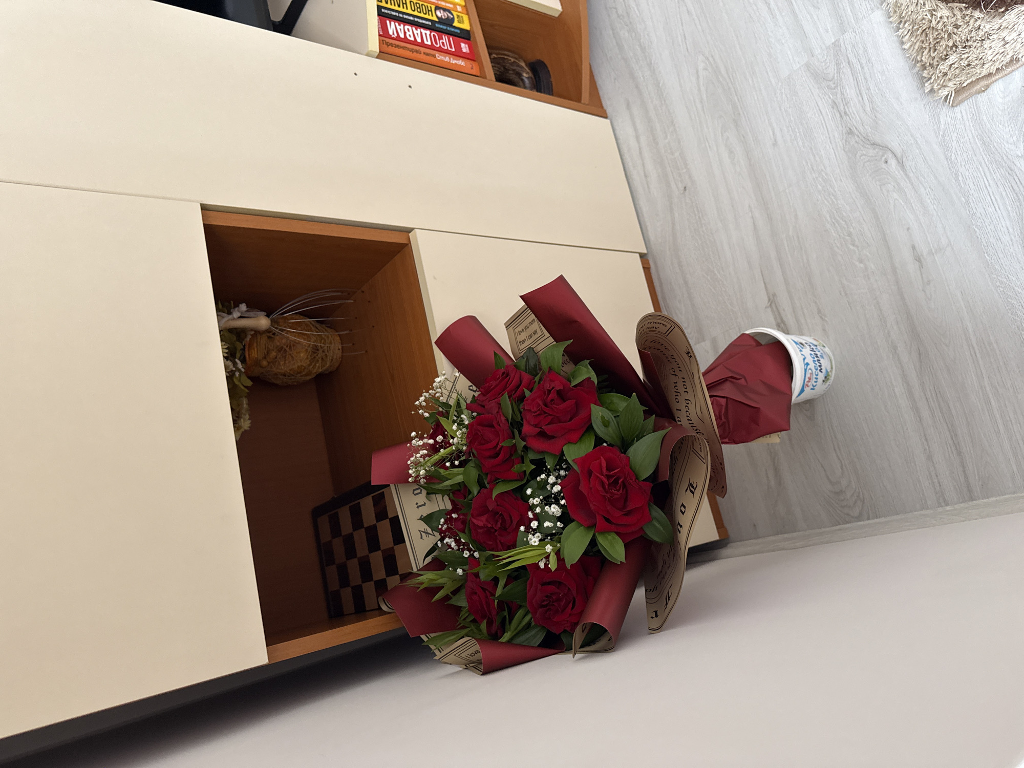 The image displays a vibrant bouquet of red roses, accompanied by sprigs of white baby's breath and green foliage, wrapped in a combination of deep red and patterned brown paper. The bouquet lies diagonally across a light-colored, possibly white, surface, with its base extending towards a light-colored wooden floor. A disposable cup, featuring Cyrillic text including "млян", is tucked into the red wrapping of the bouquet.
In the background, to the left, is a two-tiered cabinet or shelving unit with off-white doors and visible wooden shelves. On the upper shelf, several books are stacked, some with Cyrillic titles visible, such as "НОВО НАЧАЛО" (NEW BEGINNING), "ПРОДАВАЙ" (SELL), and "Презентирай като Стив Джобс" (Present like Steve Jobs). Within a lower, open compartment of the cabinet, a decorative object resembling a wire whisk with a natural fiber base, and a checkered box, are visible. The floor is made of light-toned wooden planks, and a corner of a shaggy, light-brown rug is visible in the upper right.
Visible text on the bouquet's patterned wrapping includes phrases like "I love you more then I can say", "I love you not because", "Haruse of who I a", and other fragmented words such as "dec", "mo", "Zero", and "10正". The scene captures an indoor setting, likely in a home or office, in Varna, Bulgaria, indicated by the location context and the Cyrillic text on the books and cup. No specific activity is taking place; the image is a still life.