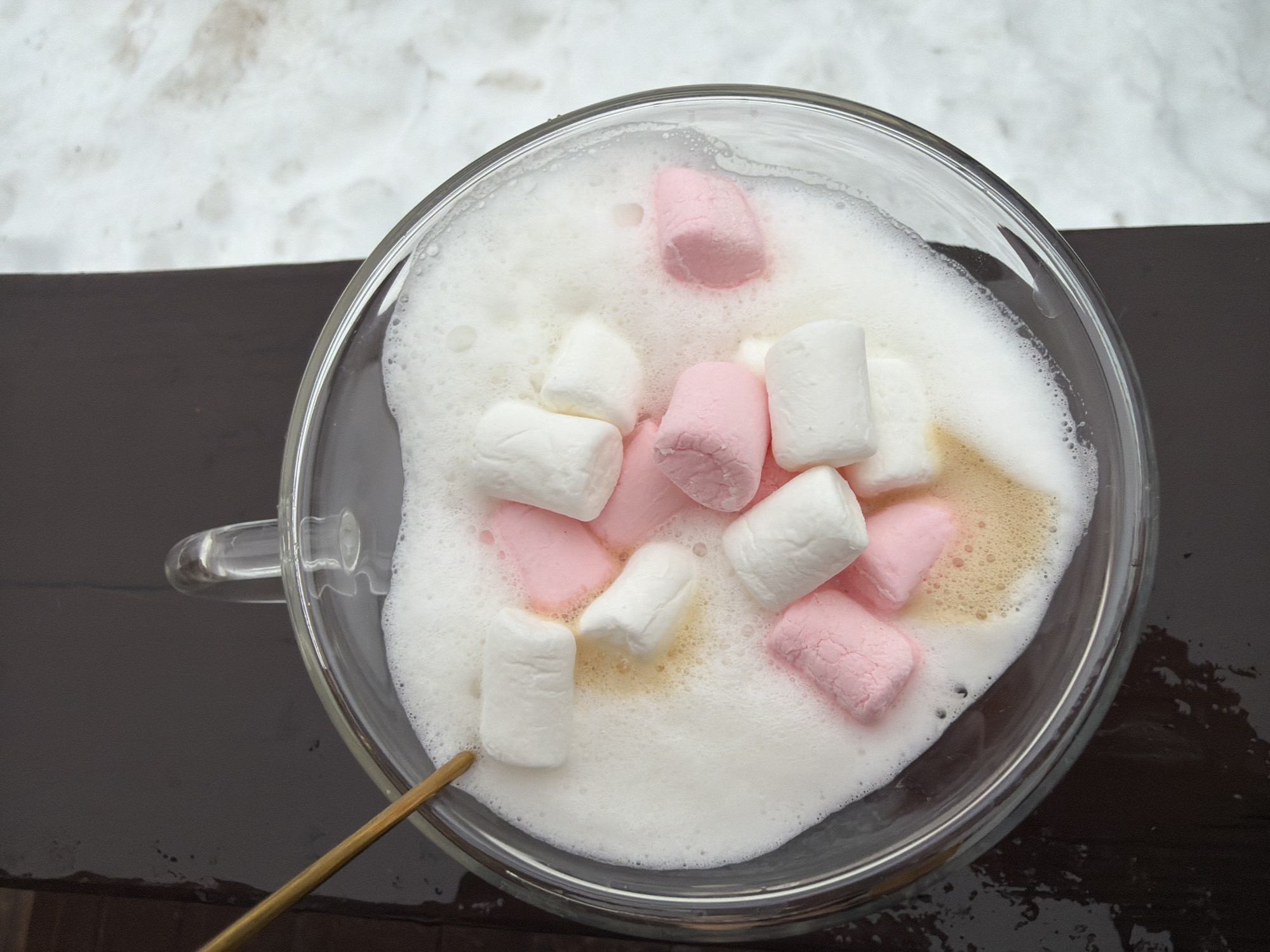 The image shows a clear glass mug filled with a white, foamy liquid, likely a beverage, with several pink and white marshmallows floating on top. A thin, light-brown stirring stick rests on the rim of the mug, partially submerged in the liquid. The mug sits on a dark brown surface, possibly a table or countertop. In the background, a patch of white snow is visible. There is no information in the image to indicate the specific location of this scene, but you specified that it is located in Chaikovsky, Russia.