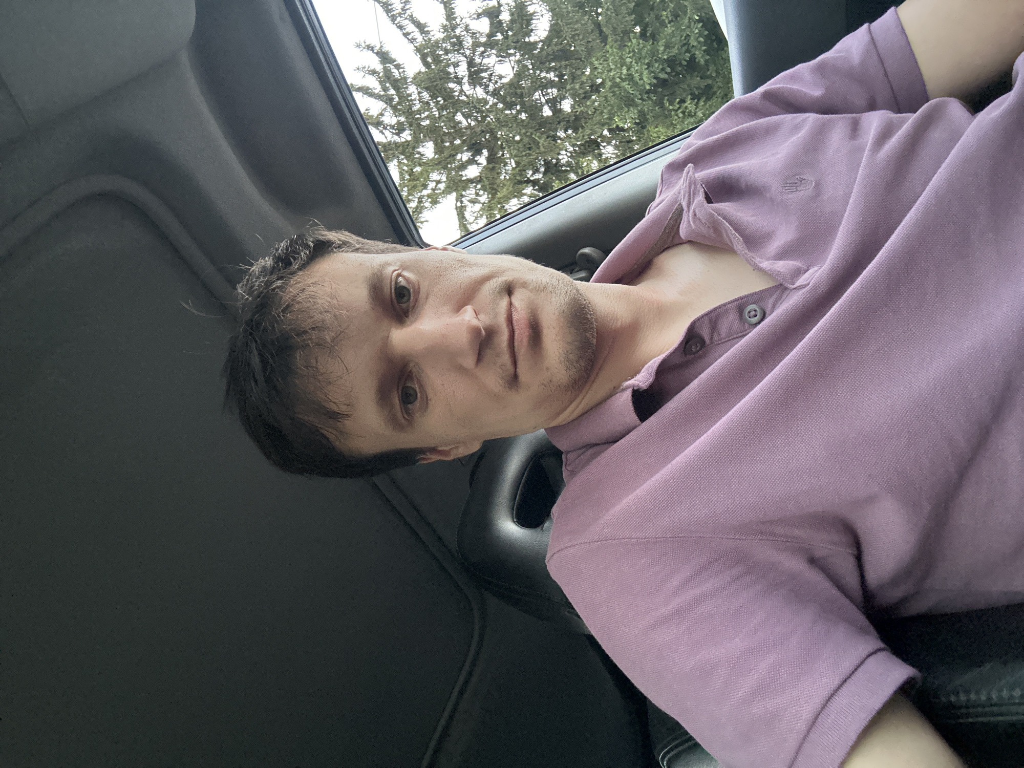 This is a selfie taken from inside a car, looking towards the driver's side. The subject is a Caucasian male with medium-length brown hair and a light stubble. He is wearing a purple polo shirt with a small embroidered emblem on the left chest. The car interior is visible, including a dark-colored headliner and a black seat. Outside the car window, a green evergreen tree is visible against a bright sky. The exact location in Shatava, Ukraine cannot be determined from the image.