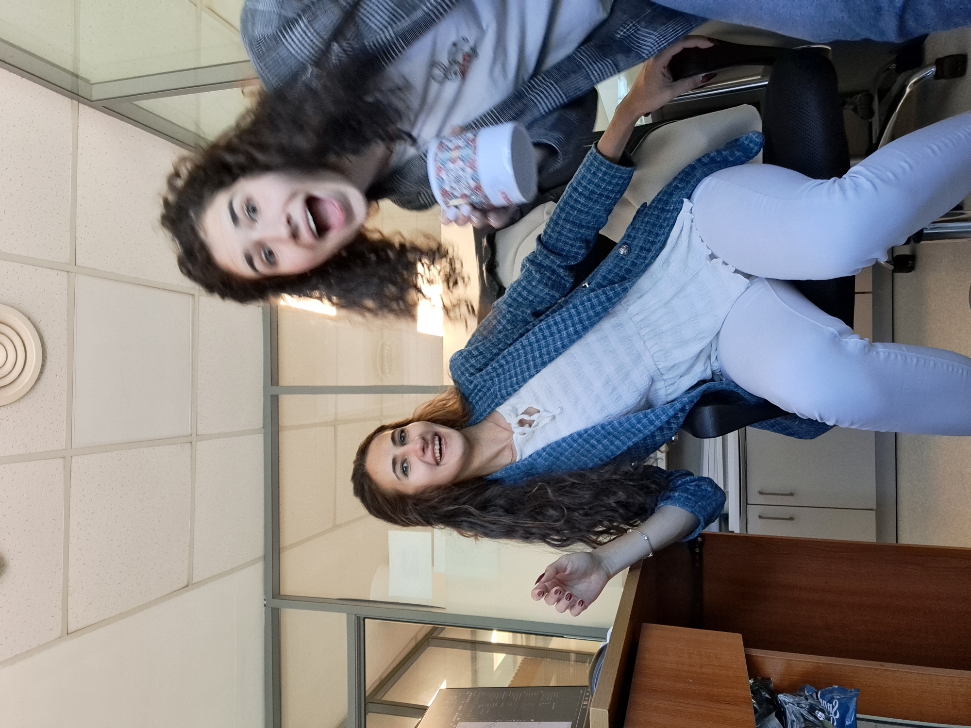Two young women are indoors, possibly in an office setting. The woman on the left is upside down in the upper portion of the frame, holding a cup and opening her mouth wide. The woman on the right is sitting in a chair, wearing a blue jacket over a white top, and white pants. The room features a windowed wall, a desk, and a drop ceiling. The location cannot be definitively confirmed as Sofia, Bulgaria without additional context.