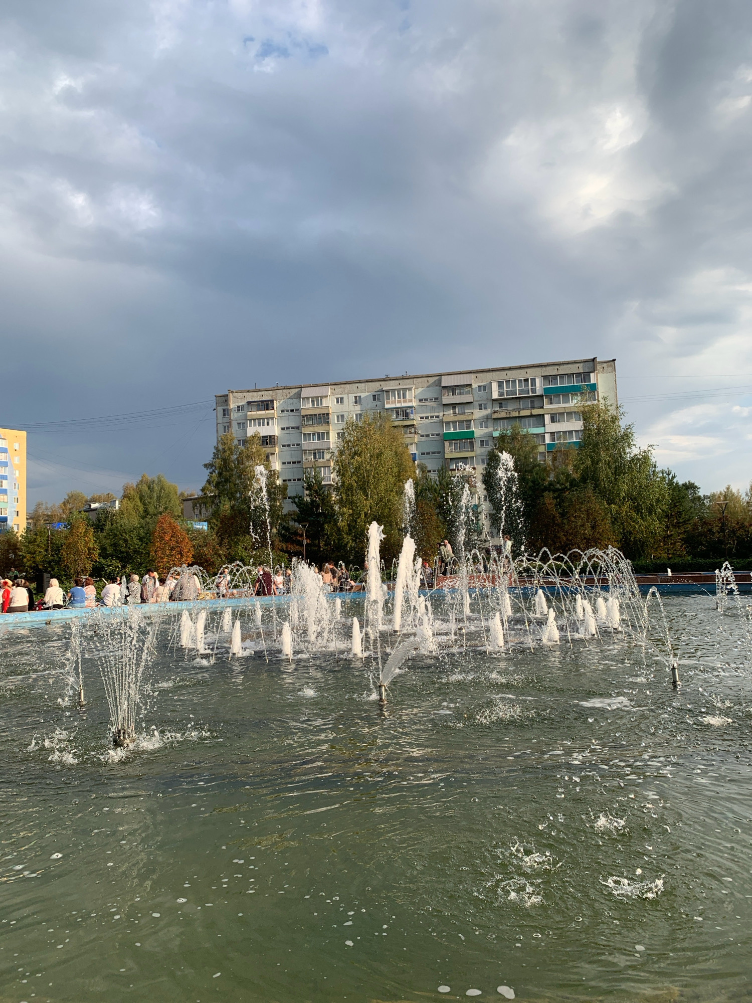 This is a medium-shot, eye-level, outdoor, daytime photograph taken in Prokopyevsk, Russia, showing a fountain in a park with a residential building in the background. The fountain is active, with multiple jets of water spraying upwards and outwards into a large, dark green body of water. In the foreground, the water is choppy with many small splashes and some foam.
Along the edge of the fountain, in the middle ground, a group of people are sitting. They appear to be casually gathered, facing the fountain. Behind the fountain and the people is a line of trees displaying autumn colors, ranging from green to yellow and orange.
Dominating the background is a tall, multi-story residential building. It is light gray with rows of windows. Some windows have balconies, and a few have green-colored frames. To the left of this building is a smaller, bright yellow residential building with white window frames. The sky above is overcast with dark, dramatic clouds, suggesting either an approaching storm or the aftermath of one. The overall atmosphere is slightly somber due to the sky, contrasted with the lively activity of the fountain and the presence of people enjoying the park.