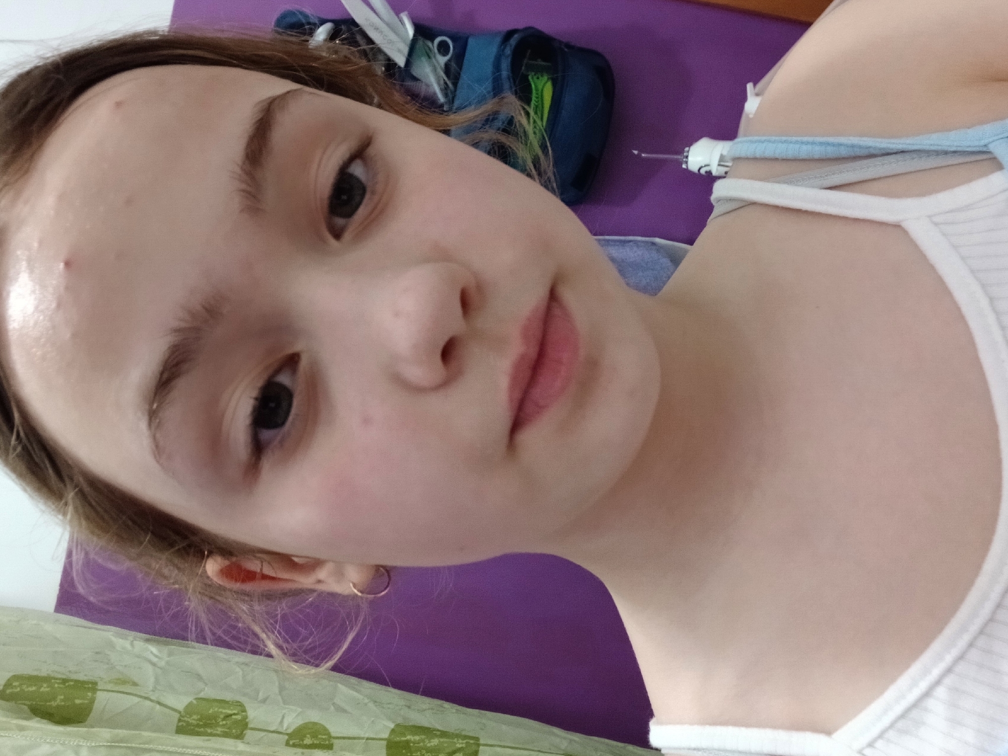A close-up, slightly angled portrait of a young female with light skin, brown hair, and brown eyes. She has a small hoop earring in her left ear. There are a few blemishes on her forehead. She is wearing a white tank top with blue straps. Behind her, there is a purple surface, a zippered blue bag with various items (scissors, green straws), and part of a green patterned object, which could be fabric or wrapping paper. The scene lacks any defining characteristics that would locate it in Krasnodar, Russia, or any specific city. The setting appears to be indoors. There is no explicit action or interaction occurring in the image. The overall impression is a casual self-portrait or snapshot.