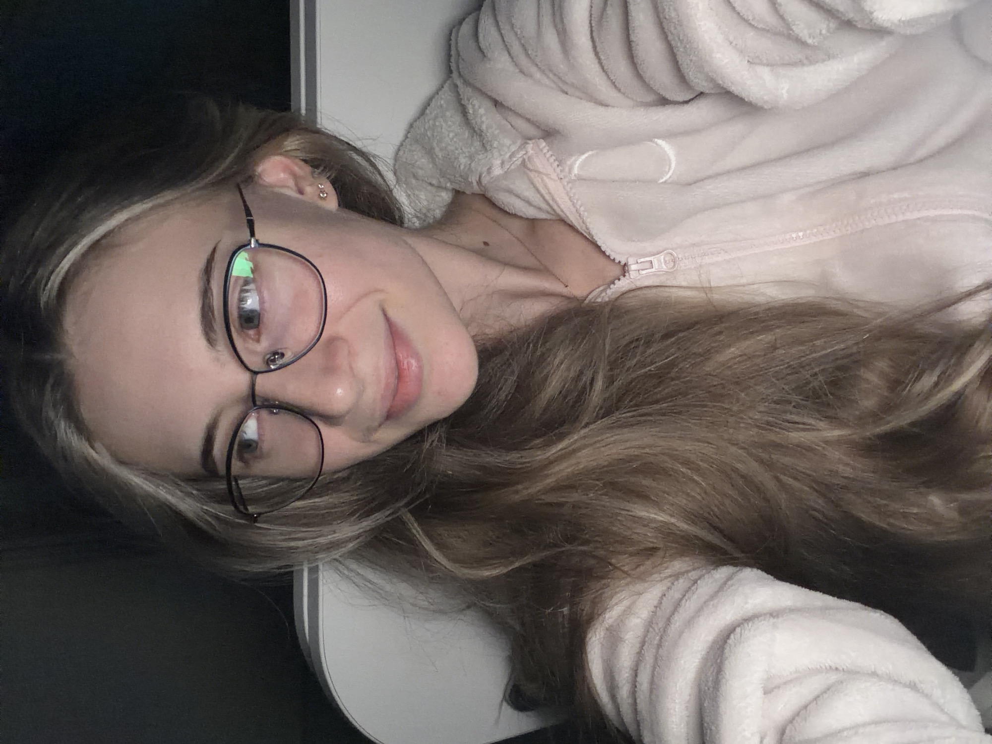 A young woman with long, blonde hair and glasses is lying down, wearing a light pink, hooded zippered jacket. She is smiling slightly at the camera. She has fair skin and two earrings in her left ear. A small silver necklace is visible at her neck. Part of a white surface is seen on the left side of the image, possibly a desk or wall. The background is dark. There is no evidence to suggest the image was taken in Plovdiv, Bulgaria.