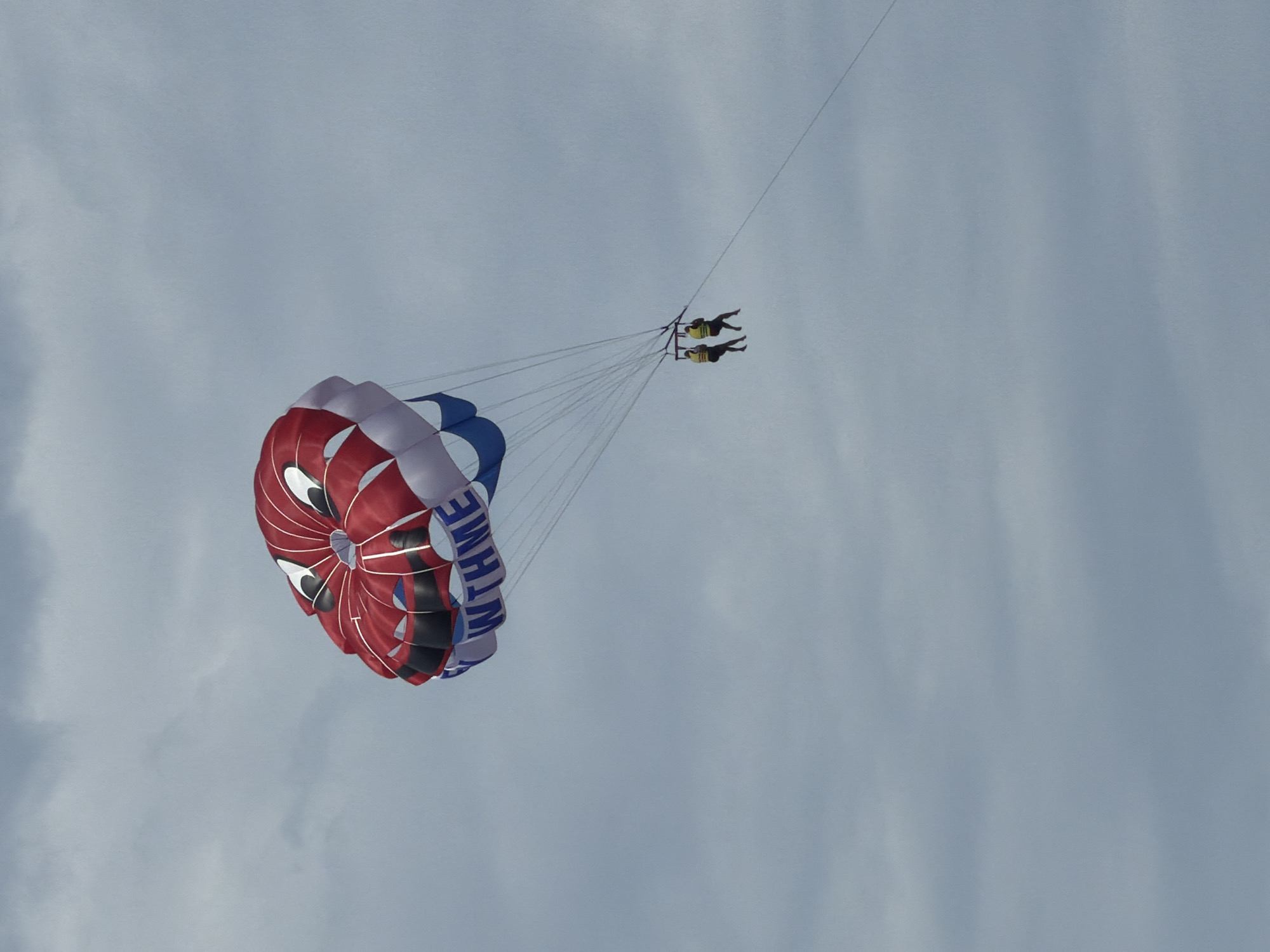 Two people are parasailing over the city of Bečići, Montenegro. They are suspended beneath a red, white, and blue parachute. The parachute is attached to a long towline that stretches upwards. The background is a cloudy sky.
