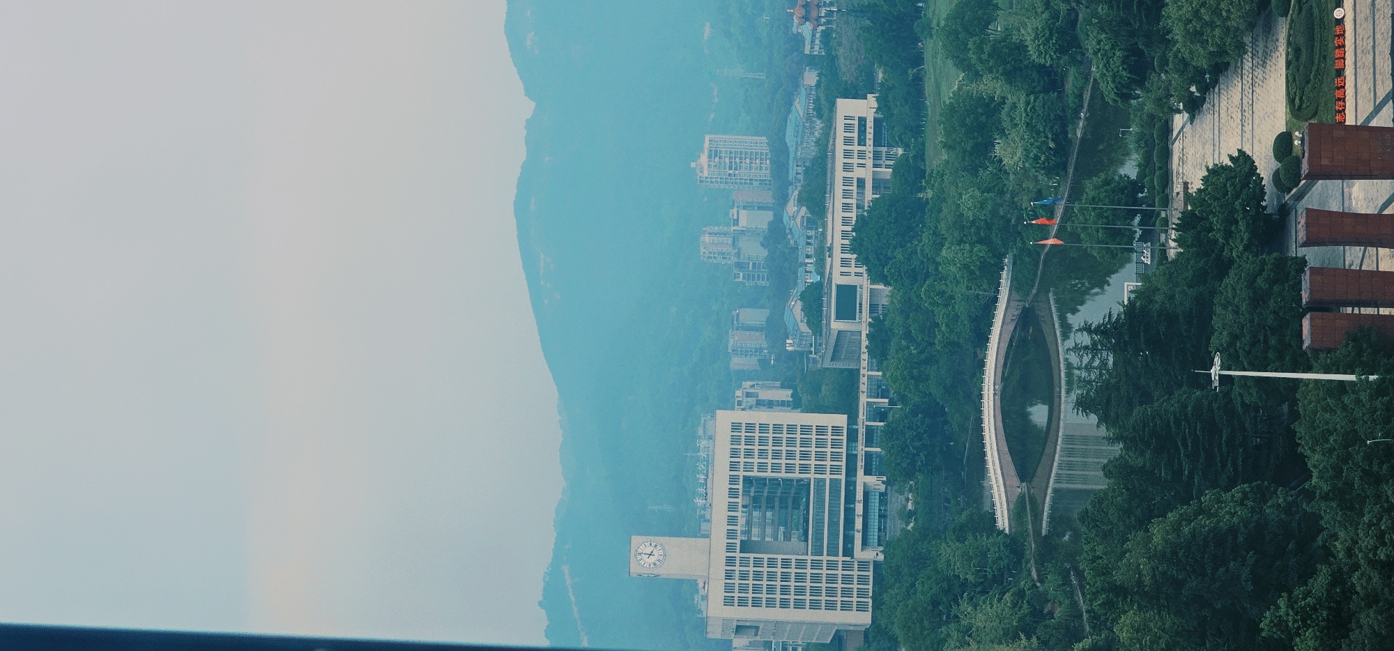 The image depicts a panoramic view of a cityscape with lush greenery and distant mountains. In the foreground, a body of water reflects the surrounding environment. A white arched bridge spans the water, leading to a paved pathway lined with trees and manicured bushes. Several flagpoles with red and blue flags stand near the water's edge.
Dominating the midground is a large, modern building complex with a prominent clock tower. This building features a distinctive facade with a large square opening and numerous windows. Adjacent to it, another long, rectangular building is visible, also with a facade of windows. The buildings appear to be part of an educational or institutional campus.
In the background, a range of blue-hued mountains forms the horizon. Scattered amongst the trees and closer to the mountains are several taller, contemporary buildings, suggesting a developed urban area. The sky is overcast with a pale, hazy light.
The location is identified as the city of Chongqing High-tech Zone, China. The scene suggests a well-maintained campus or park area integrated with urban development. No people are discernible in the image. The overall impression is one of a serene, well-ordered environment within a larger urban setting.