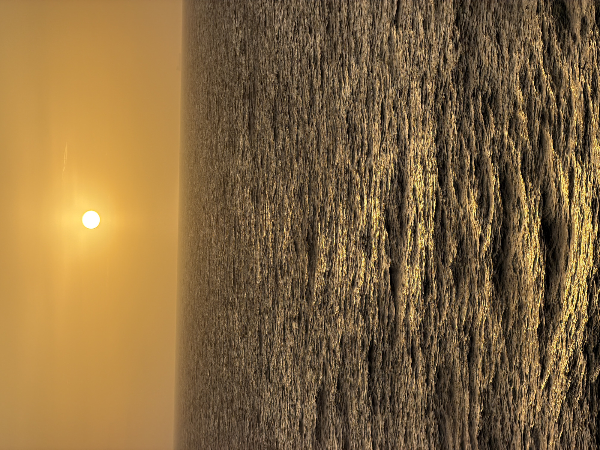 A digitally rendered image depicts a stark contrast between light and texture. On the left, a hazy, yellow-toned sky contains a bright, spherical light source, presumably the sun. A faint streak cuts across the sky above the sun. To the right, a textured, vertical surface with irregular, striated patterns extends from top to bottom. The lighting accentuates the texture, creating a sense of depth. The overall atmosphere is surreal and dreamlike. The location details are unavailable.