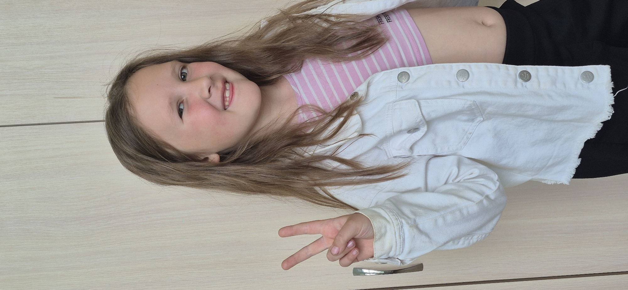 A young girl lies on her side with her head tilted towards the viewer, against what appears to be a light-colored wooden surface. She is making a peace sign with her right hand. She has long brown hair and is wearing a pink and white striped crop top, over which is a white denim jacket. She also appears to be wearing black bottoms.