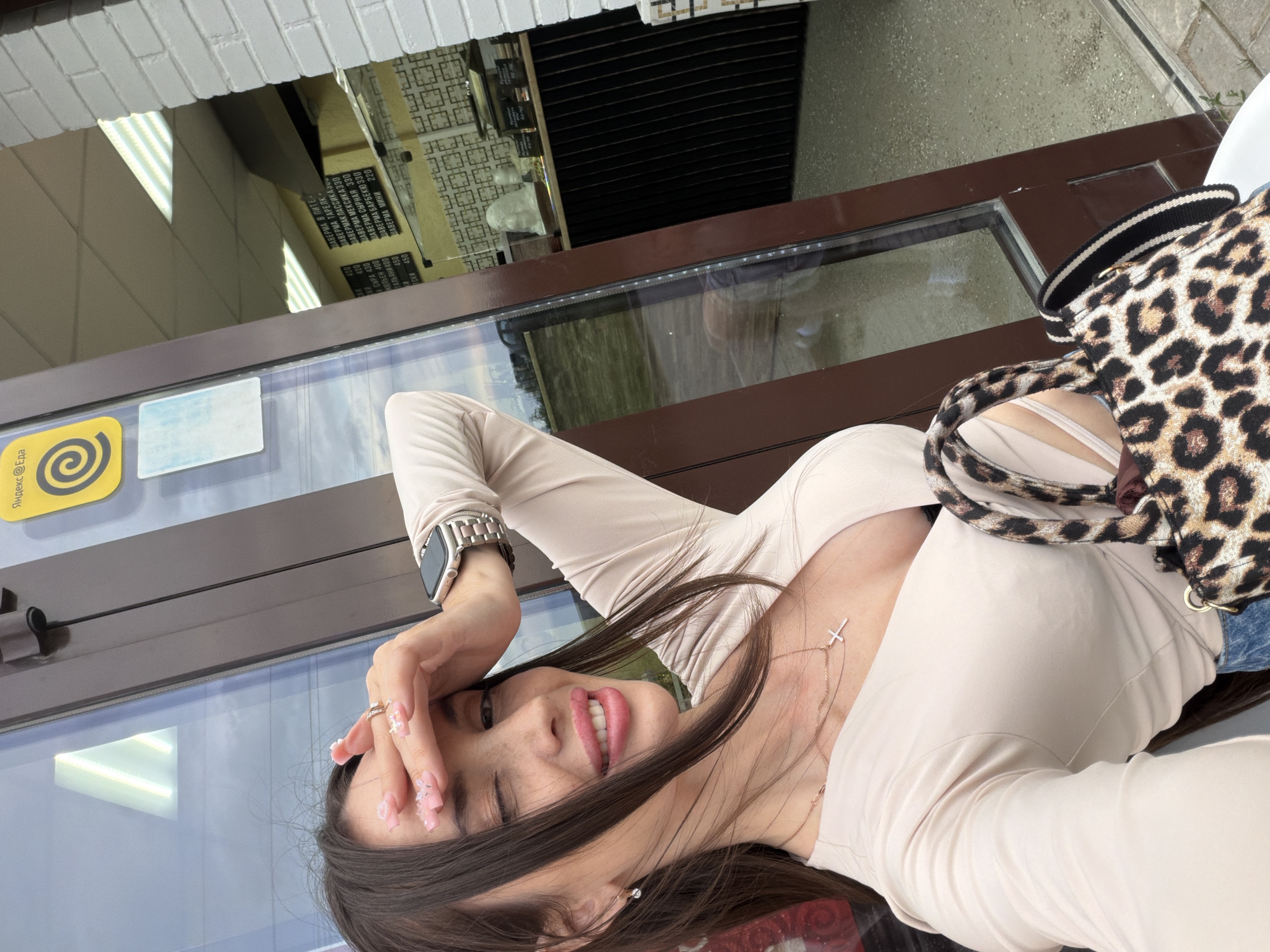 A woman takes a close-up selfie in front of a commercial establishment in Saint Petersburg, Russia, identifiable by the "Яндекс Еда" (Yandex Eda) sign and Cyrillic menu. She has long dark brown hair, a fair complexion with freckles, and wears light pink lipstick, winking with her right eye and smiling. Her left hand is raised to her forehead, shielding her eye, revealing long pink acrylic nails with white embellishments and a silver-colored ring. She wears a small diamond stud earring in her left ear, a delicate rose gold cross necklace, and a silver-colored Apple Watch on her left wrist. She is dressed in a light beige/cream long-sleeved top with a deep V-neckline. She carries a leopard-print shoulder bag.
The background shows the glass entrance/window of a food establishment. Through the glass, a dark counter with a decorative white object is visible, along with interior walls featuring a yellow base color and a repeating geometric pattern. A black menu board in Cyrillic is prominent, listing various "Шаверма" (Shawarma) options, confirming it as a shawarma eatery or fast-food cafe. Prices are listed in rubles (e.g., 420, 400, 390, 330, 220). The exterior features a white brick wall to the right and a paved ground surface. Reflections in the glass show the sky and a distant landscape, suggesting an outdoor setting.