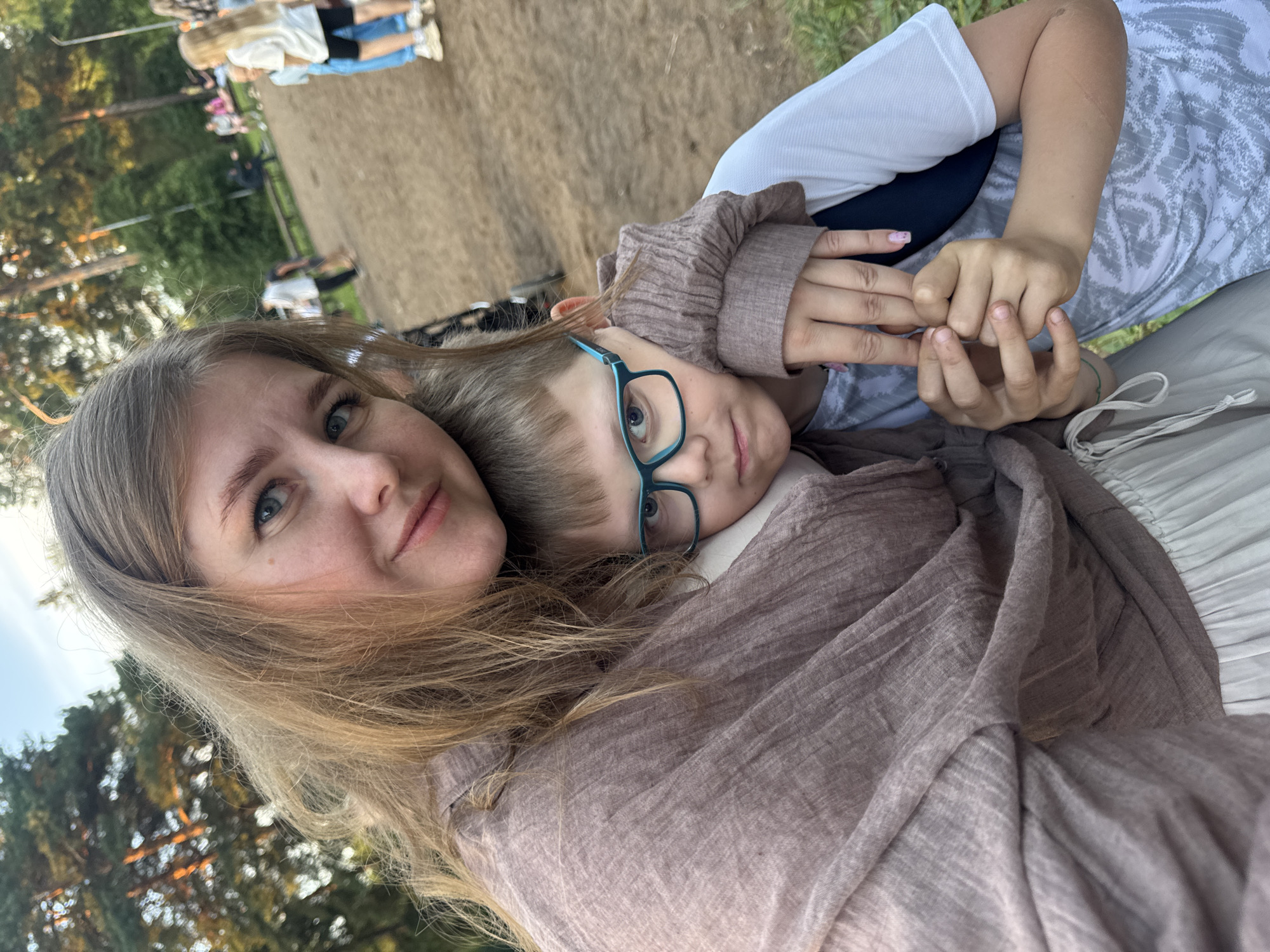 A mother and son are posing for a selfie outdoors in a park-like setting in Горный, Russia. The mother, in the foreground on the left, is wearing a light brown, textured shawl and has long blonde hair. She is holding the son, who is on the right, closer to her. The son is wearing glasses with blue frames and a white t-shirt with a gray pattern. He is looking slightly to the side, with his arm around his mother's shoulder. In the background, other people are visible, some sitting on benches and others walking. The ground appears to be a dirt path, and trees are visible in the distance. The lighting suggests it is late afternoon or early evening.