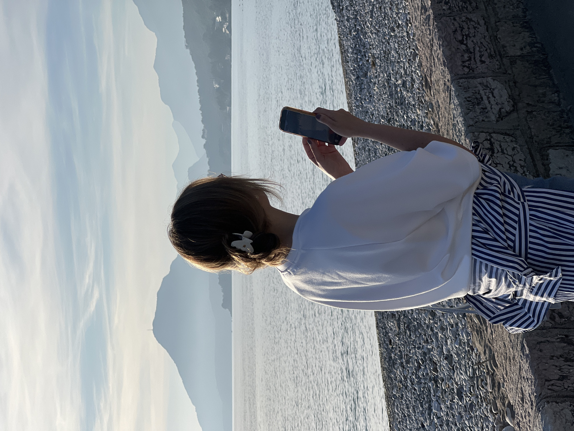 An outdoor, sunlit image captures a person from behind, standing by a body of water and holding a smartphone horizontally with both hands, seemingly photographing the scenic view. The person has short brown hair styled in a low bun with a white floral clip on the right side. They are wearing a loose-fitting white t-shirt and what appears to be striped blue and white bottoms, possibly a skirt or wide-leg trousers, tied at the waist.
The scene depicts a vast expanse of water, likely the sea or a large lake, stretching into the distance under a bright sky. In the background, multiple layers of mountains with varying shades of blue and gray rise, suggesting depth and distance. The sky above is light blue with faint, wispy clouds. The lighting indicates late afternoon or early morning, with warm sunlight catching the person's hair and clothing, as well as glinting off the water's surface.
To the right of the person, a textured pathway or embankment made of small, light-colored stones or pebbles runs parallel to the water's edge. Further to the right, a darker, rougher stone wall or structure is visible. On the distant left shore, small, white structures can be faintly seen, suggesting distant habitation. The setting is consistent with a coastal or lakeside location like Bar, Montenegro. No visible text can be clearly discerned on the smartphone screen or elsewhere.