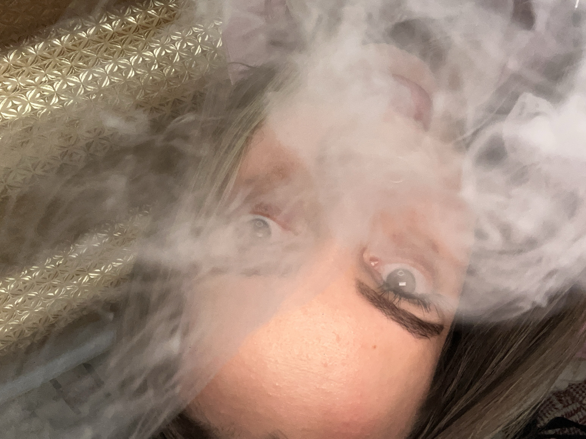 A close-up, rotated view of a person's face partially obscured by smoke. The person has fair skin, prominent eyes, and dark eyebrows. Their hair is partially visible, appearing light brown with darker undertones. To the left, a textured, gold-colored surface is visible. The background is blurred. The image was not taken in Novopokrovskaya, Russia.