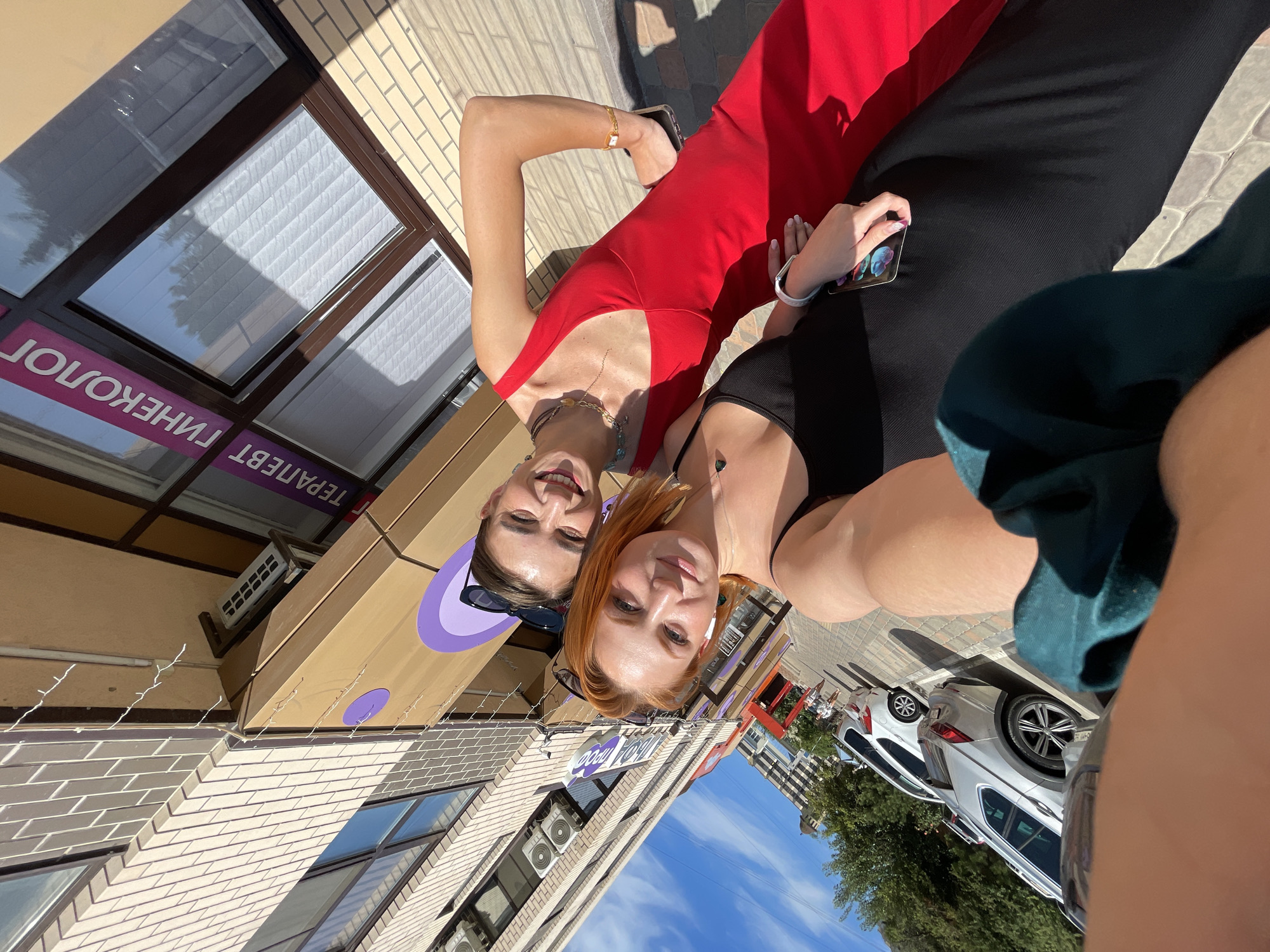 Two women are taking a selfie outdoors. The woman in the foreground has bright red hair and is wearing a black dress. The woman behind her has dark hair and is wearing a bright red dress. They are standing in front of a building with a sign that reads "ТЕРАПЕВТ ГИНЕКОЛОГ" (Therapist Gynecologist). The building appears to be a medical clinic or office. The background shows a street with cars parked and trees. The sky is blue with some clouds. The overall setting is urban. The location is the city of Anapa, Russia.