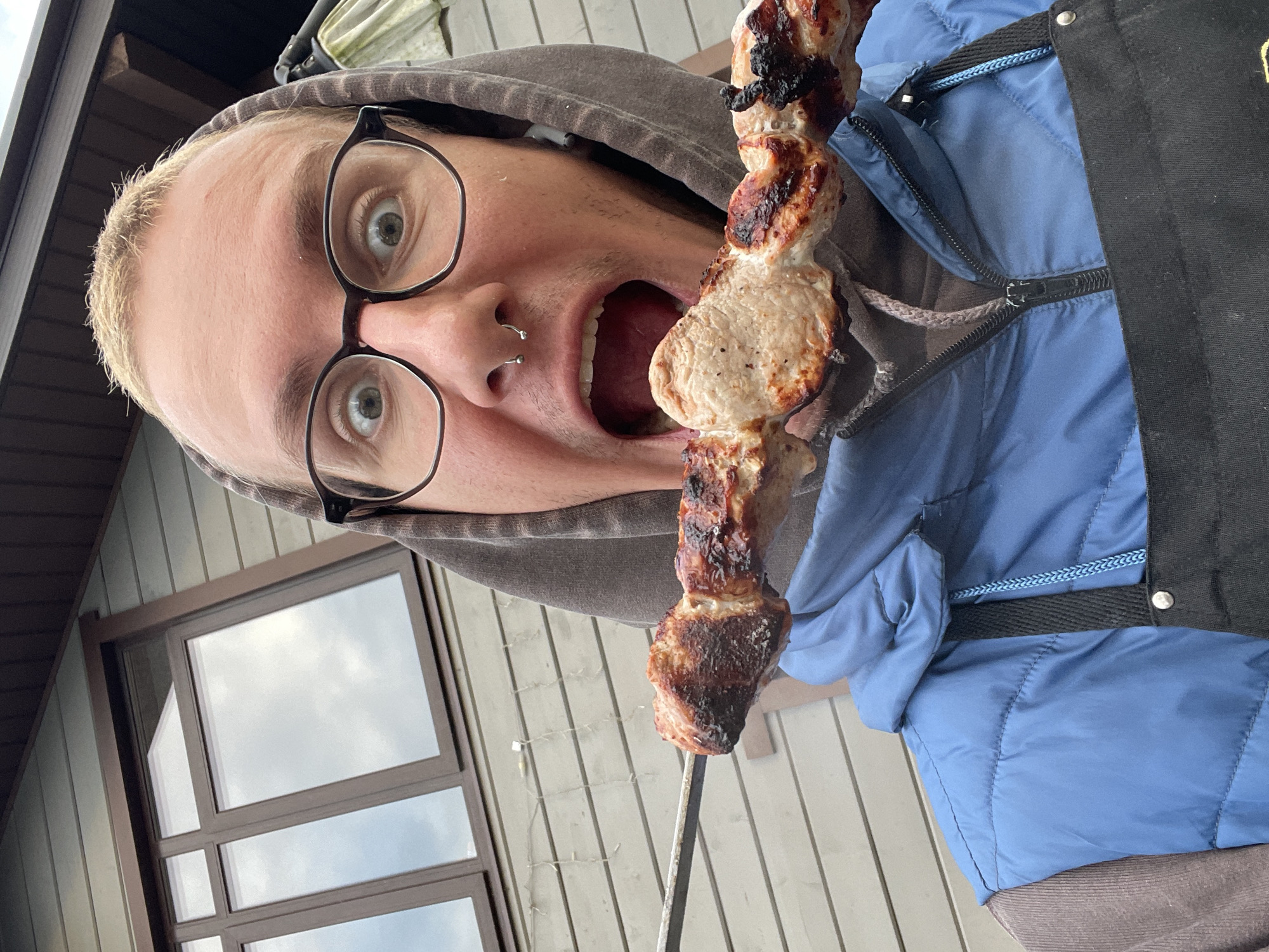A person wearing glasses, a gray hooded sweatshirt, and a blue quilted jacket with a black apron is holding a metal skewer with cooked meat in front of their open mouth. The person has a nose ring and is looking directly at the camera with wide eyes. The background shows a light-colored wooden building with a window. The location is plausibly Sestroretsk, Russia, considering the architectural style.