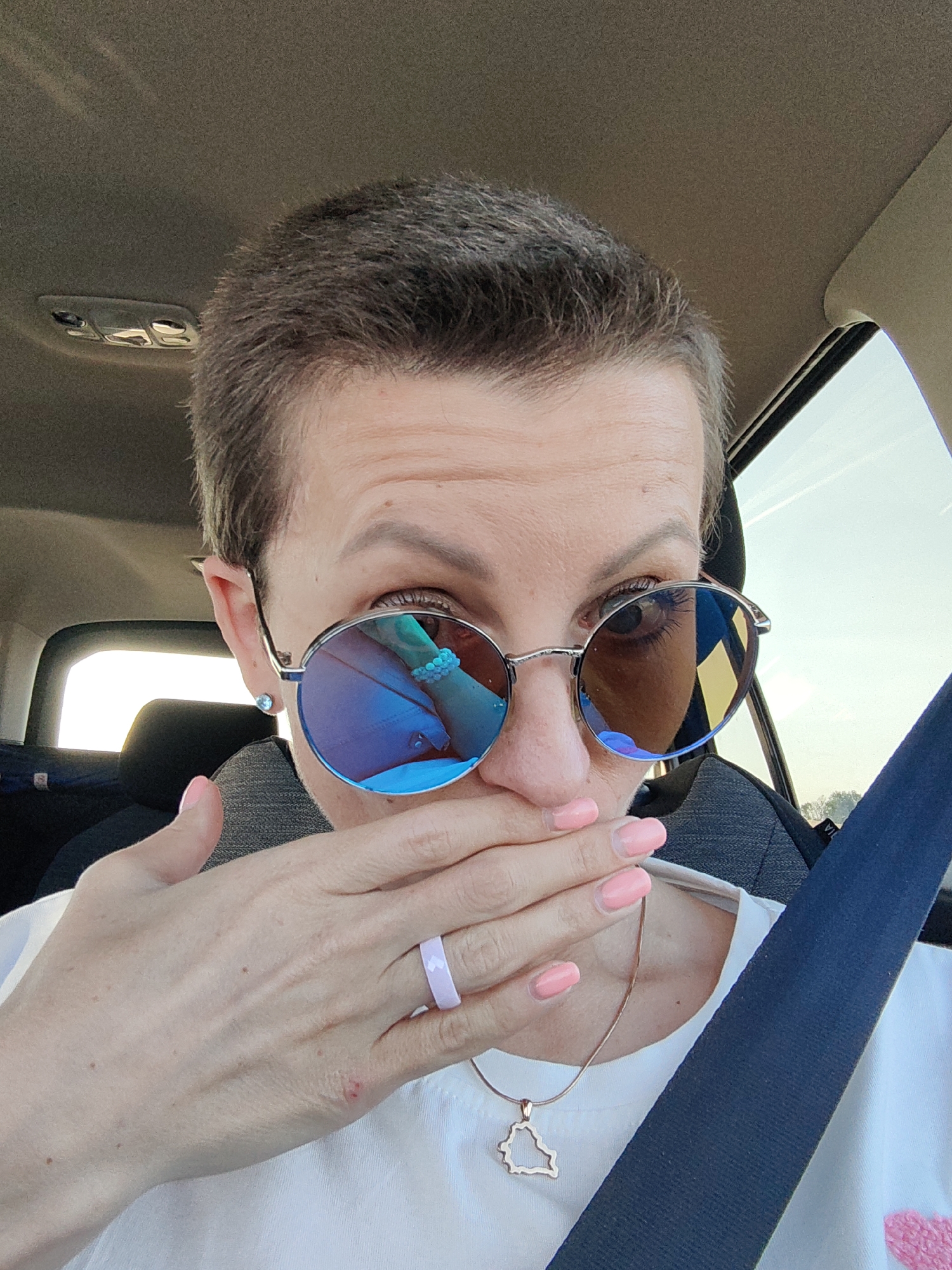 The image shows a close-up, selfie-style shot of a person with short hair, wearing sunglasses and a white t-shirt, inside a vehicle. The person is covering their mouth with their right hand. Their fingernails are painted a light pink color, and they are wearing a light purple ring on their left ring finger and a delicate necklace with a pendant shaped like a country. The sunglasses have blue-tinted lenses that reflect the interior and exterior of the car, as well as the person's arm and jewelry. The background shows part of the car's interior, including the ceiling, a headrest, and a seatbelt. The exterior view through the car window suggests an outdoor environment with some sky visible. The image does not provide any specific visual cues to determine the precise location within Міханавіцкі сельскі Савет, Belarus.