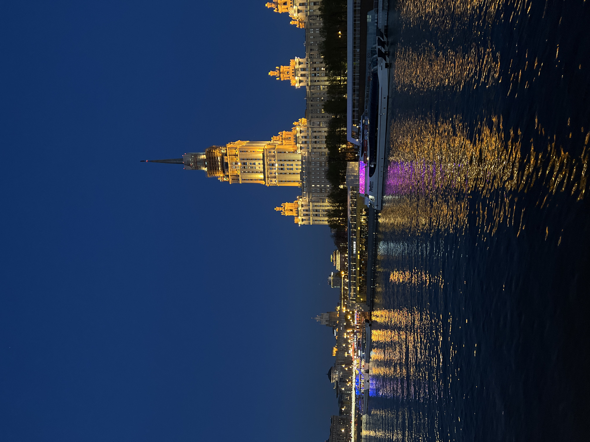 The image depicts a night scene in Moscow, Russia, specifically showcasing the illuminated Kotelnicheskaya Embankment Building reflected in the Moskva River. A boat with purple lighting is docked near the embankment. The dark blue sky is above the building. The reflections of the lights in the water create golden streaks.