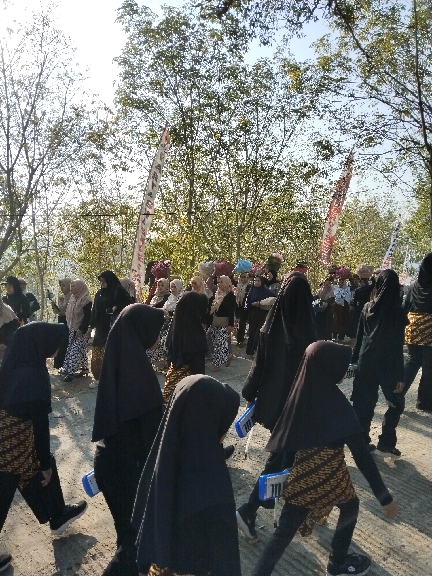 In Musuk, Indonesia, a group of women is participating in a parade, with some carrying baskets of produce on their heads. They are dressed in traditional attire, including hijabs and batik skirts. The parade is taking place outdoors, with trees and a clear sky in the background. The women are walking on a dirt road, and some of them are holding musical instruments, possibly melodicas. The overall atmosphere appears to be festive and celebratory.