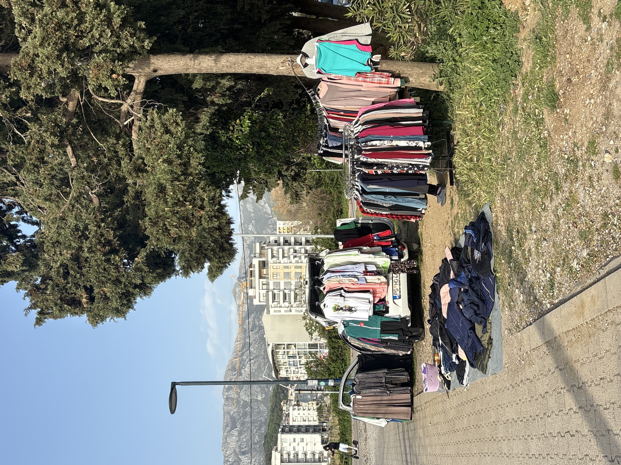 The image depicts a clothing sale set up on a sidewalk in the city of Bar, Montenegro. A white car with license plate "PG•HG442" is parked adjacent to the sidewalk, with a substantial amount of clothing hanging on racks and piled on a blanket. The clothing appears to be a mix of styles and colors. In the background are several multi-story buildings, mountains, and a construction crane. A street lamp is visible. The sky is clear. There is a large tree to the left of the clothing display, and grassy area. A person is seen riding a scooter down the street.