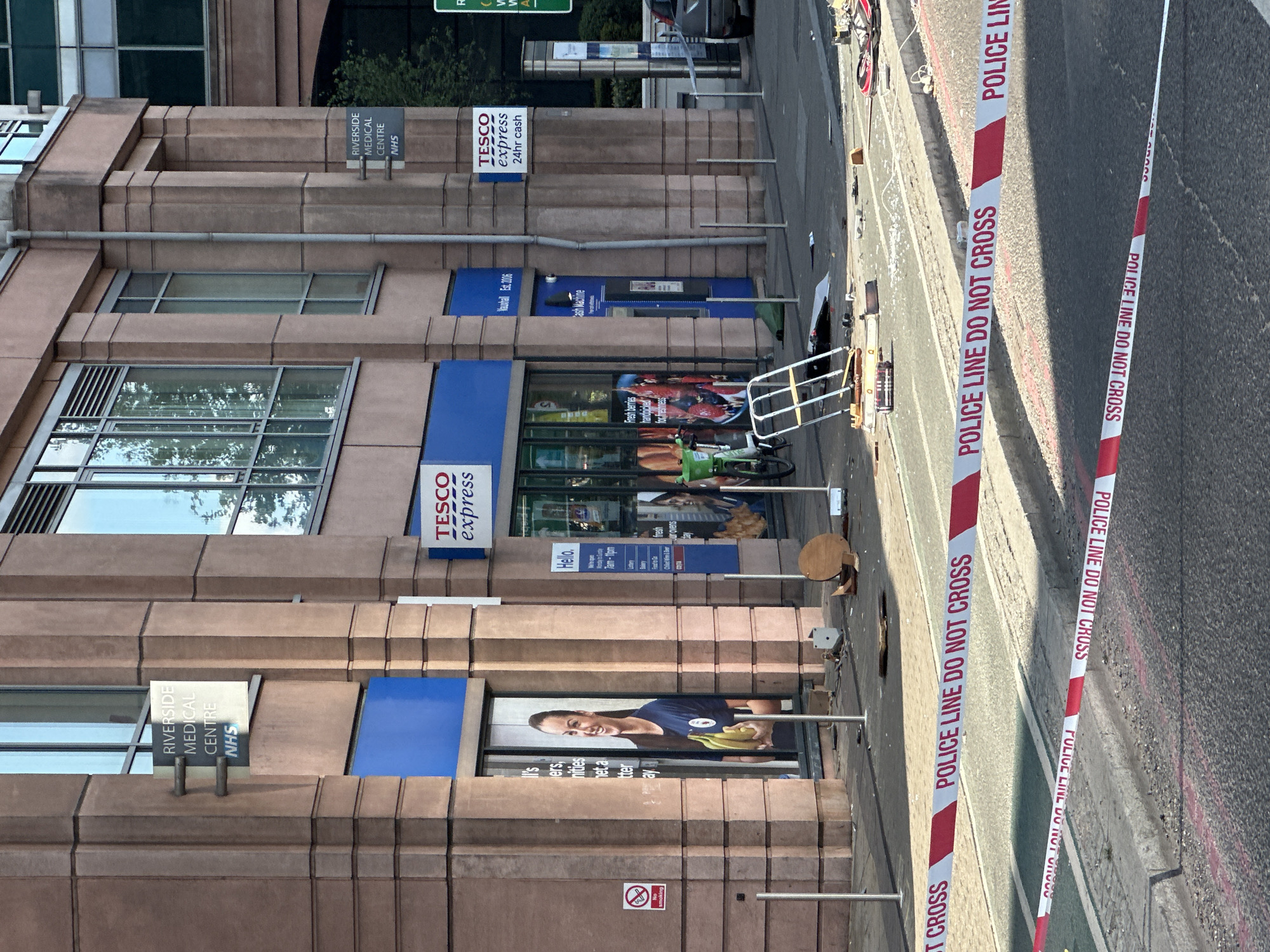 The image shows the exterior of a Tesco Express store located next to Riverside Medical Centre NHS in what appears to be a city environment. The Tesco Express features standard signage, including a poster of a woman holding bananas and information about opening hours. An ATM is also present. The scene is cordoned off with "POLICE LINE DO NOT CROSS" tape. The immediate area includes a sidewalk and a street. Other miscellaneous objects are visible, including a bike and street furniture.