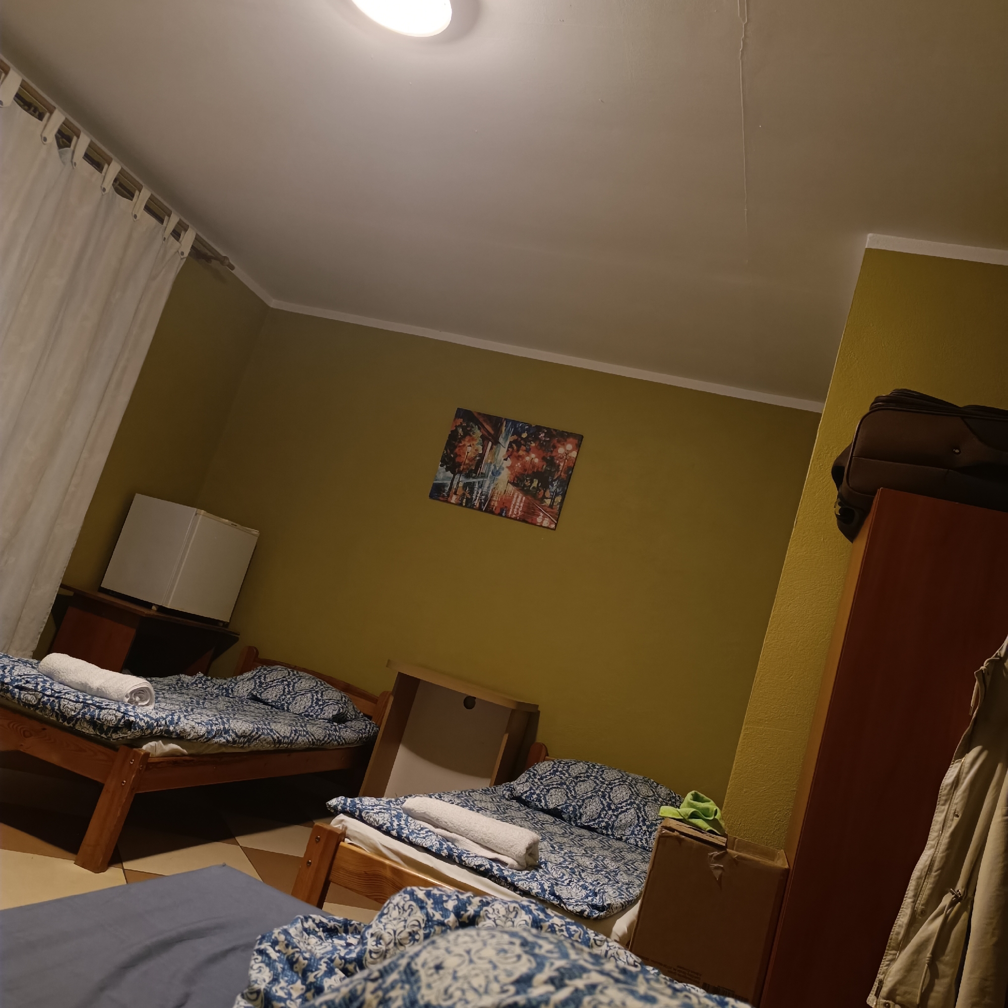 This is a photograph of a room in Mielno, Poland, that appears to be a hostel or shared accommodation. The room contains multiple beds with blue and white patterned bedding. On one of the beds, a white towel is neatly folded. A small white refrigerator sits on a dark wooden desk or cabinet. A piece of artwork depicting a street scene at night hangs on the yellow wall. A dark brown suitcase is stacked on top of a tall, reddish-brown wardrobe or cabinet. A beige jacket hangs from the side of the wardrobe. A cardboard box with a green item on top is placed near the beds. The room is illuminated by an overhead light, casting shadows. The floor is tiled in a diamond pattern. Curtains are visible on the left side of the image.