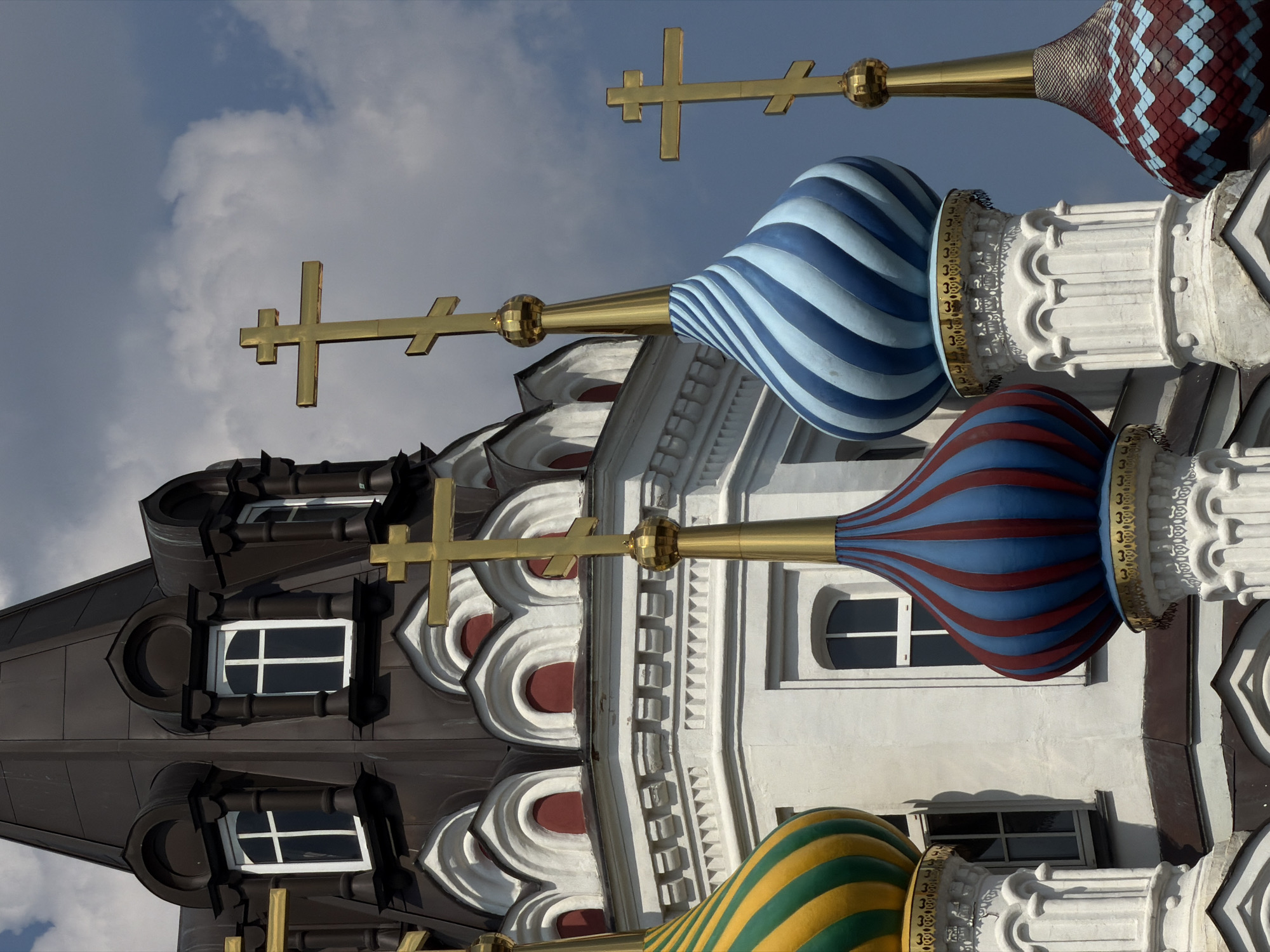 The image captures a close-up view of several onion domes of the Holy Trinity Cathedral in Saratov, Russia. The cathedral, with its distinctive Russian Orthodox architecture, features a dark gray, conical roof topped with arched dormer windows. Several golden crosses adorn the peaks of the domes. The domes themselves display a variety of colorful patterns: one is striped green and yellow, another is striped blue and white, and a third is striped red and blue. A fourth dome, partially visible on the right, is covered in a red and blue scale-like pattern. The white, ornate bases of the domes are visible, supported by columns. The sky above is a mix of blue and white clouds.