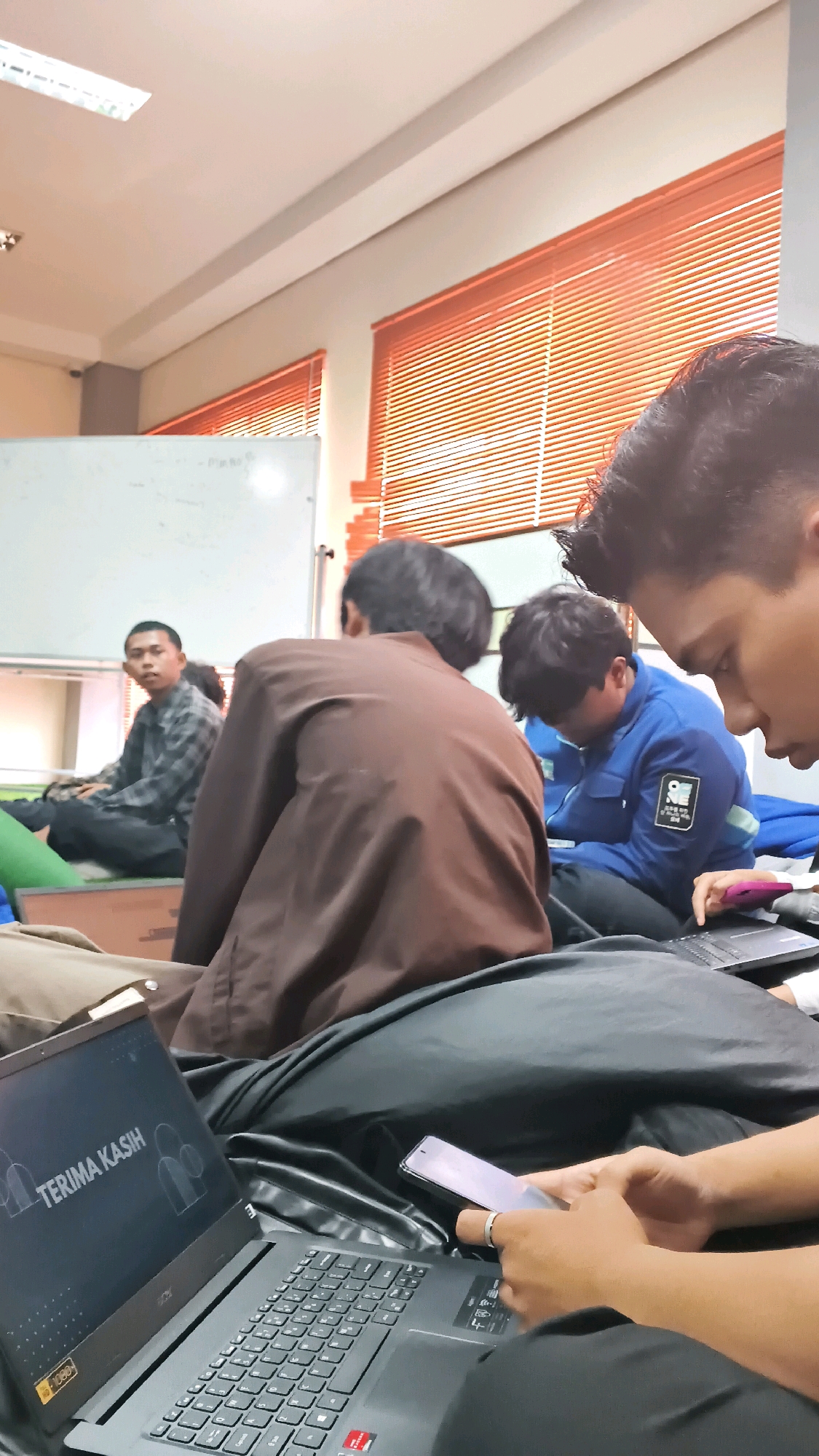 The image shows a group of young men in what appears to be a classroom or meeting room. The room has whiteboards, blinds on the windows, and fluorescent lighting. In the foreground, a laptop is open and displaying the text "TERIMA KASIH". A person's hands are visible holding a smartphone and resting on the laptop. In the middle ground, three young men are seated. One man, wearing a brown jacket, is facing away from the camera. Another man, wearing a blue jacket with patches, is looking down at something in his lap. A third man, wearing a checkered shirt, is looking towards the camera. The overall scene suggests a casual gathering or a break in a study session. The image does not provide any specific visual cues to pinpoint the location to Lohbener, Indonesia.