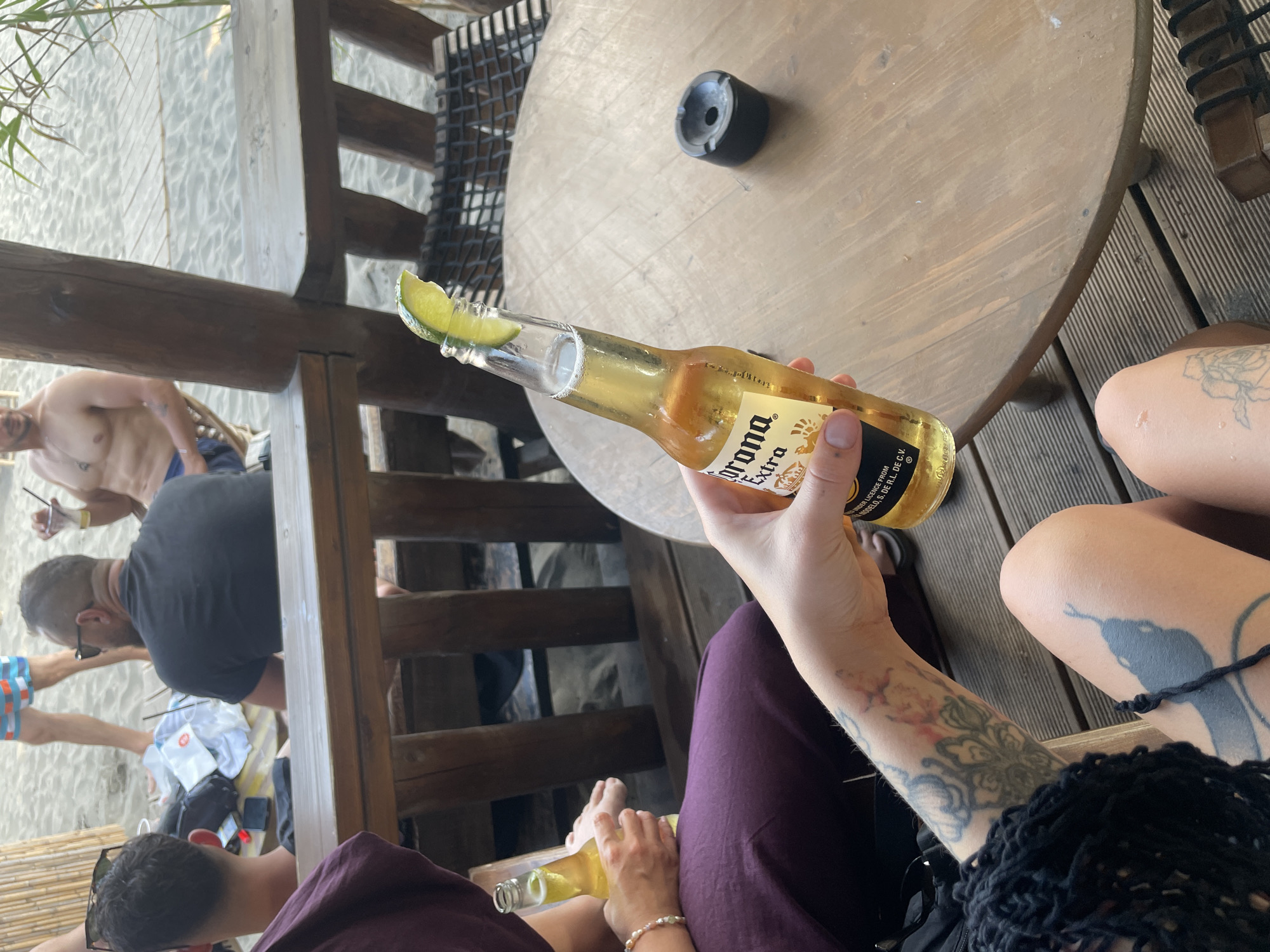 A group of people are gathered on a beach in Donji Štoj, Montenegro. The scene appears to be a beach bar or restaurant, with wooden tables and chairs set up on a sandy surface. In the foreground, a person's hand holds a bottle of Corona Extra beer, complete with a lime wedge. The person's tattooed arm is visible, along with part of their leg also featuring tattoos. In the background, other individuals are present, some seated and others standing. The atmosphere suggests a relaxed, casual setting, likely a daytime social gathering.
