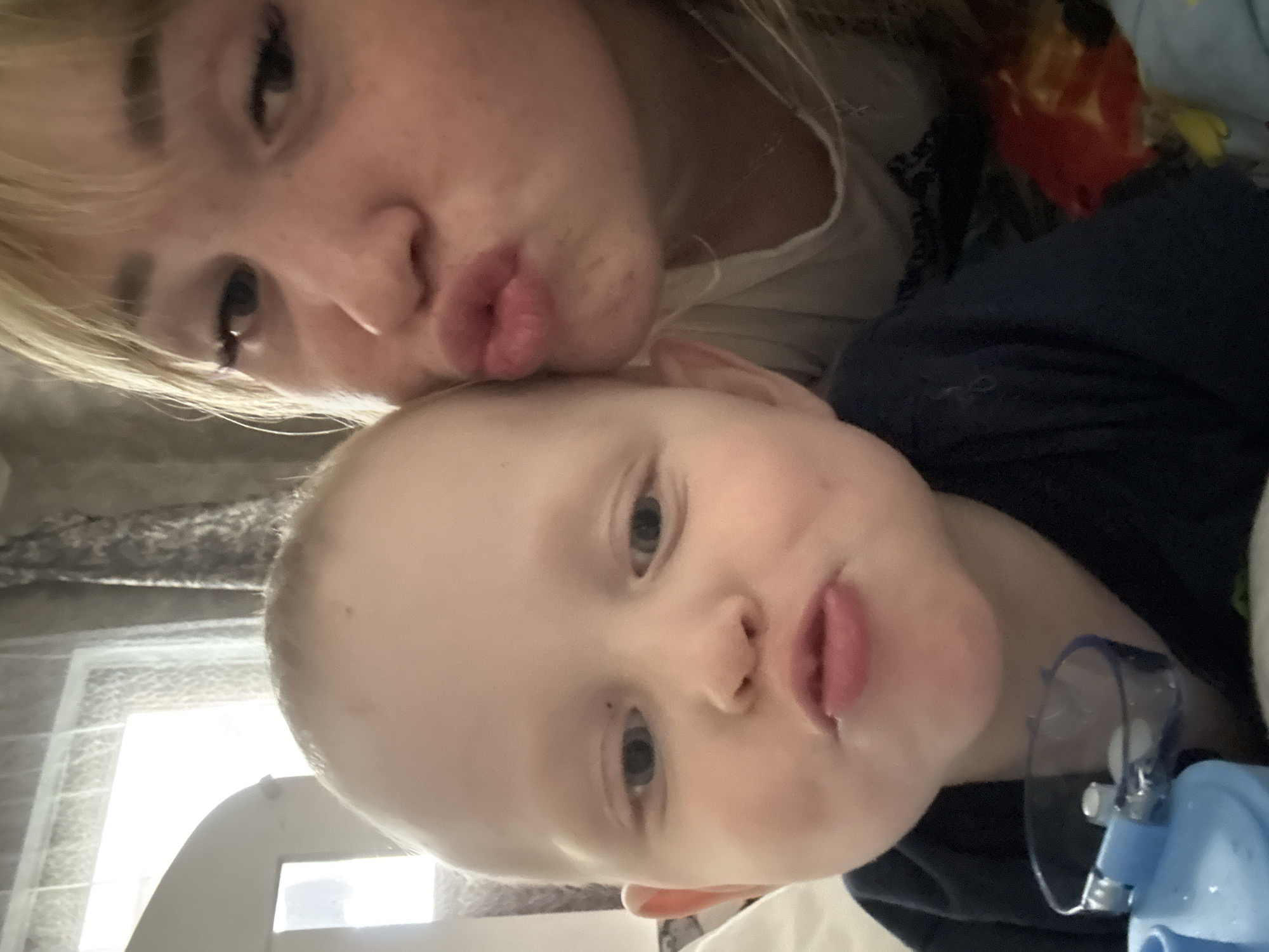 A selfie taken indoors features an adult and a young child. The adult, likely a woman, is positioned in the upper left of the frame with her face close to the camera, puckering her lips as if for a kiss. She has fair skin, blonde hair with bangs, and visible eyelashes. The child, possibly a toddler, is in the lower part of the frame. The child also puckers their lips. A piece of blue medical equipment is also visible. The background details are minimal, revealing a curtained window and what appears to be the edge of a bed. Марковский, Russia is not discernable in the image.