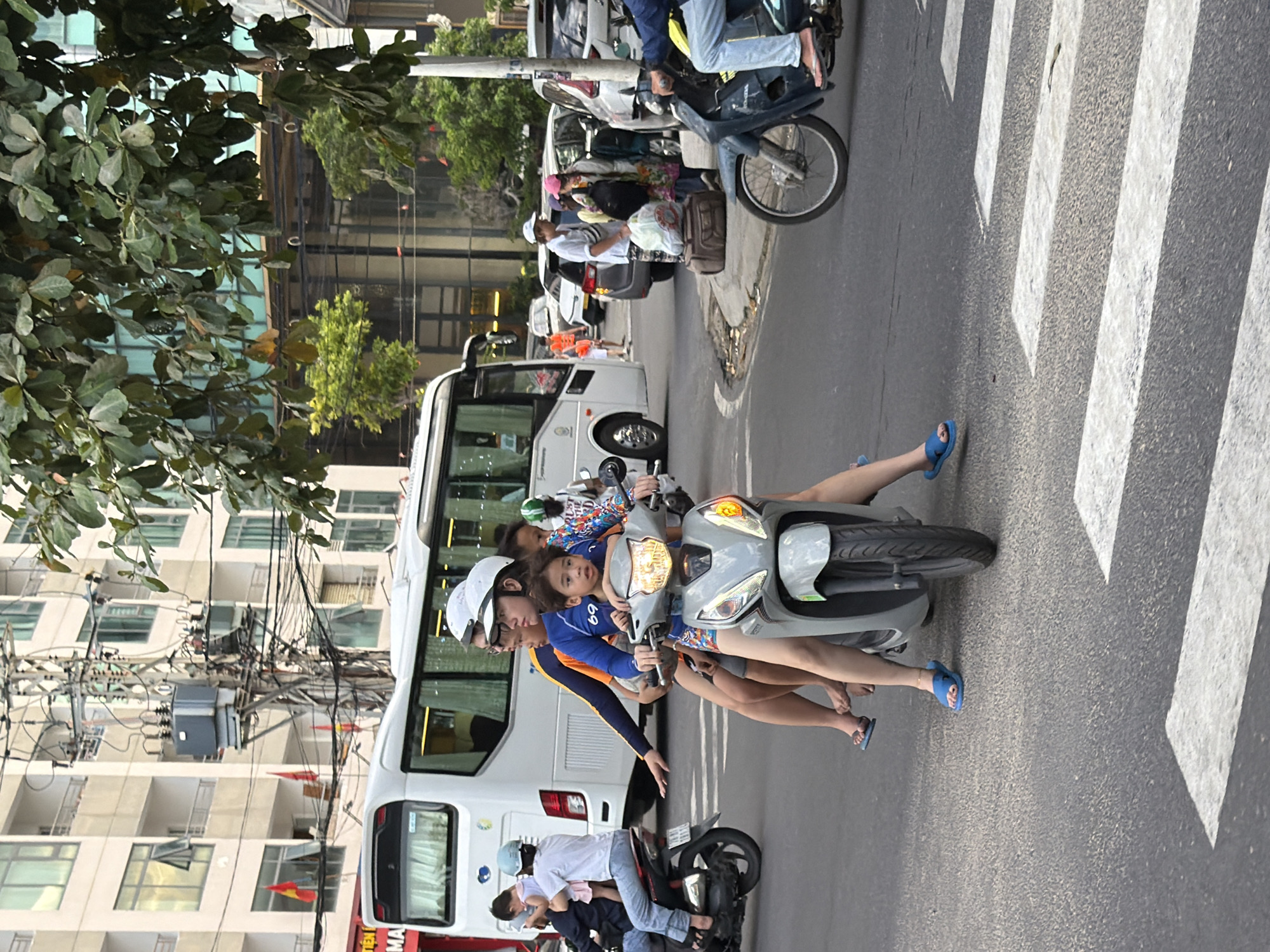 A family of four is riding a scooter on a street in Nha Trang, Vietnam. The scooter is grey and the family is dressed in casual clothing. The father is driving the scooter, and the mother is sitting behind him with a young child in her lap. Another child is sitting in front of the father. In the background, a white bus and several cars are visible. The street is paved with asphalt, and there is a pedestrian crossing in the foreground. The buildings in the background are multi-story structures with balconies and windows. Trees and foliage are also present, suggesting a tropical climate. The overall scene depicts a typical urban environment in Vietnam.