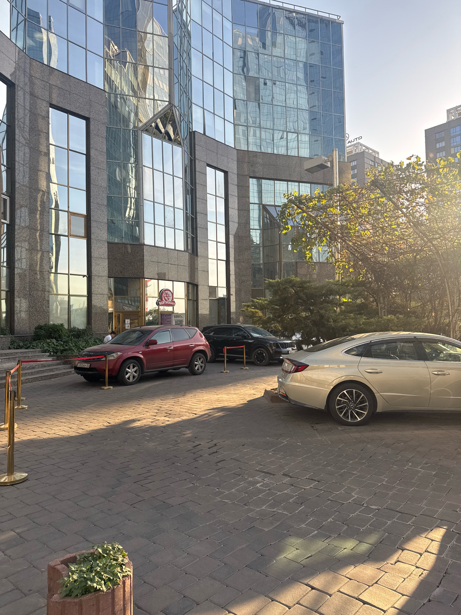 The image depicts the exterior of the "Red Dragon" restaurant, located at 129 Zhibek Zholy Street in Almaty, Kazakhstan. The building features a modern glass facade that reflects the surrounding cityscape and sky. In front of the restaurant, several cars are parked on a cobblestone pavement. A red SUV is positioned closest to the entrance, with a black SUV parked behind it. Further down, a beige sedan is visible. Red velvet ropes on stanchions demarcate a pathway leading to the building's entrance. Lush greenery, including bushes and trees, is present to the left and right of the scene. The lighting suggests it is late afternoon, with warm sunlight casting long shadows on the ground. The sky is clear and blue.