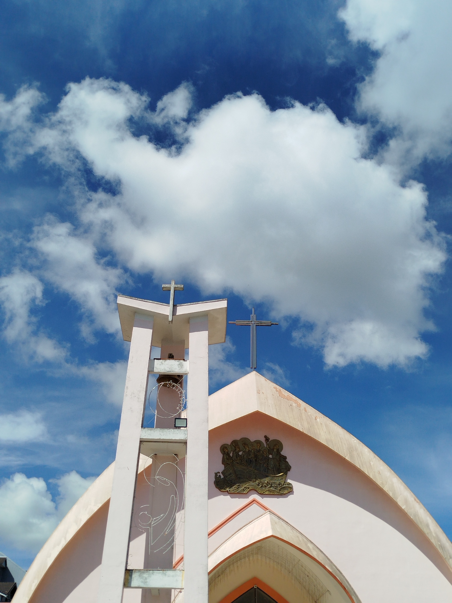 A low-angle shot of a church in Da Lat, Vietnam, set against a bright blue sky with scattered white clouds. The building is light pink with two visible crosses atop different structures. The first cross, made of grey concrete, sits on a bell tower that also features a decorative wire sculpture and a bell-shaped object. The second cross, constructed from shiny metal, is atop a peaked roof above a bas-relief depicting figures in a boat. The architectural style is modern, with curved edges and geometric shapes. The surrounding environment is only partially visible, with a glimpse of dark-colored roofing in the lower left corner.