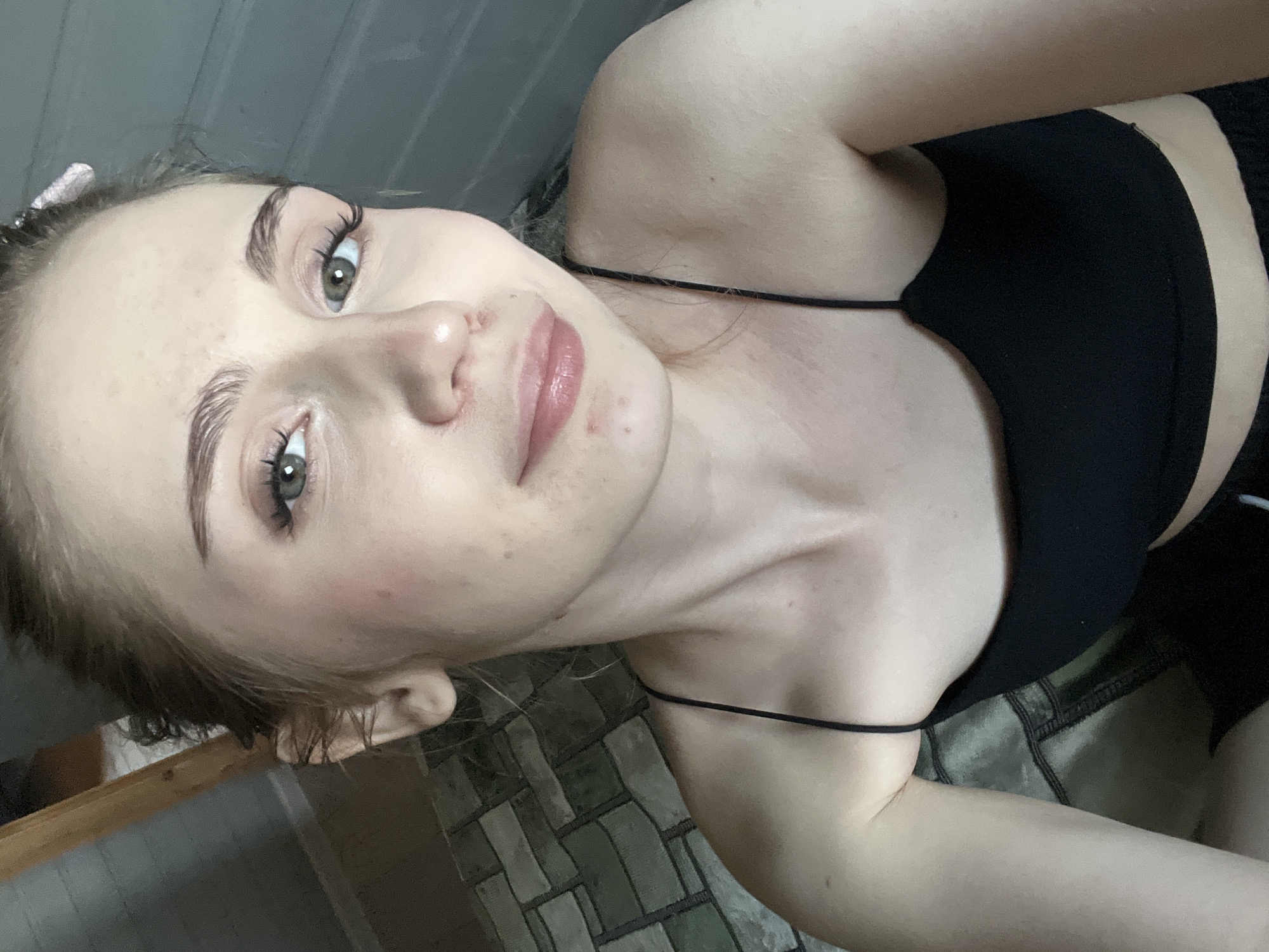 This is a medium close-up, selfie-style photograph of a young woman looking towards the camera. She has fair skin, light brown hair pulled back, and light green eyes. Her eyebrows are groomed and defined, and she is wearing subtle makeup, including mascara and lipstick. There are a few small blemishes visible on her face, particularly around her nose and chin. She is wearing a black spaghetti strap tank top. The background appears to be an indoor setting with a wall made of horizontal grey planks on the right and a textured surface resembling brick or tile on the left. A wooden beam is also visible in the upper left corner. There is no specific information in the image to indicate the location as Ashkadar, Russia.