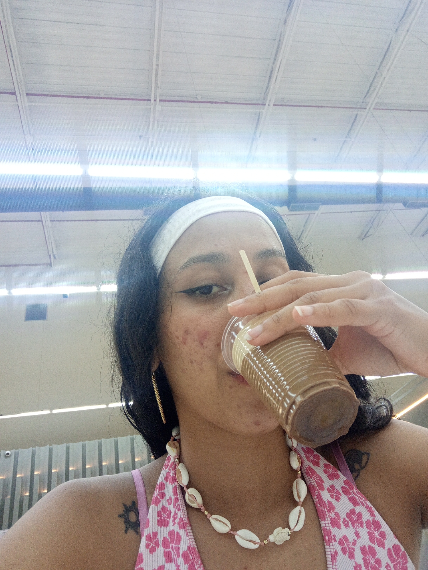 A close-up, eye-level shot features a young person, likely female, drinking from a clear plastic cup containing a light brown beverage with a straw. The person has dark hair, styled with a white headband, and is wearing winged eyeliner. Visible acne blemishes are present on their face. They are dressed in a pink and white floral patterned top, possibly a bikini top, and wear a necklace made of cowrie shells with a small turtle charm. A golden, twisted hoop earring is visible on their left ear, and a stylized sun tattoo is seen on their left shoulder. The background shows a brightly lit indoor space with a white ceiling, exposed structural beams, and long fluorescent strip lights. A thin red pipe runs along the ceiling. The setting appears to be a large commercial building, such as a shopping mall or supermarket, in Porlamar, Venezuela. No text is visible in the image.