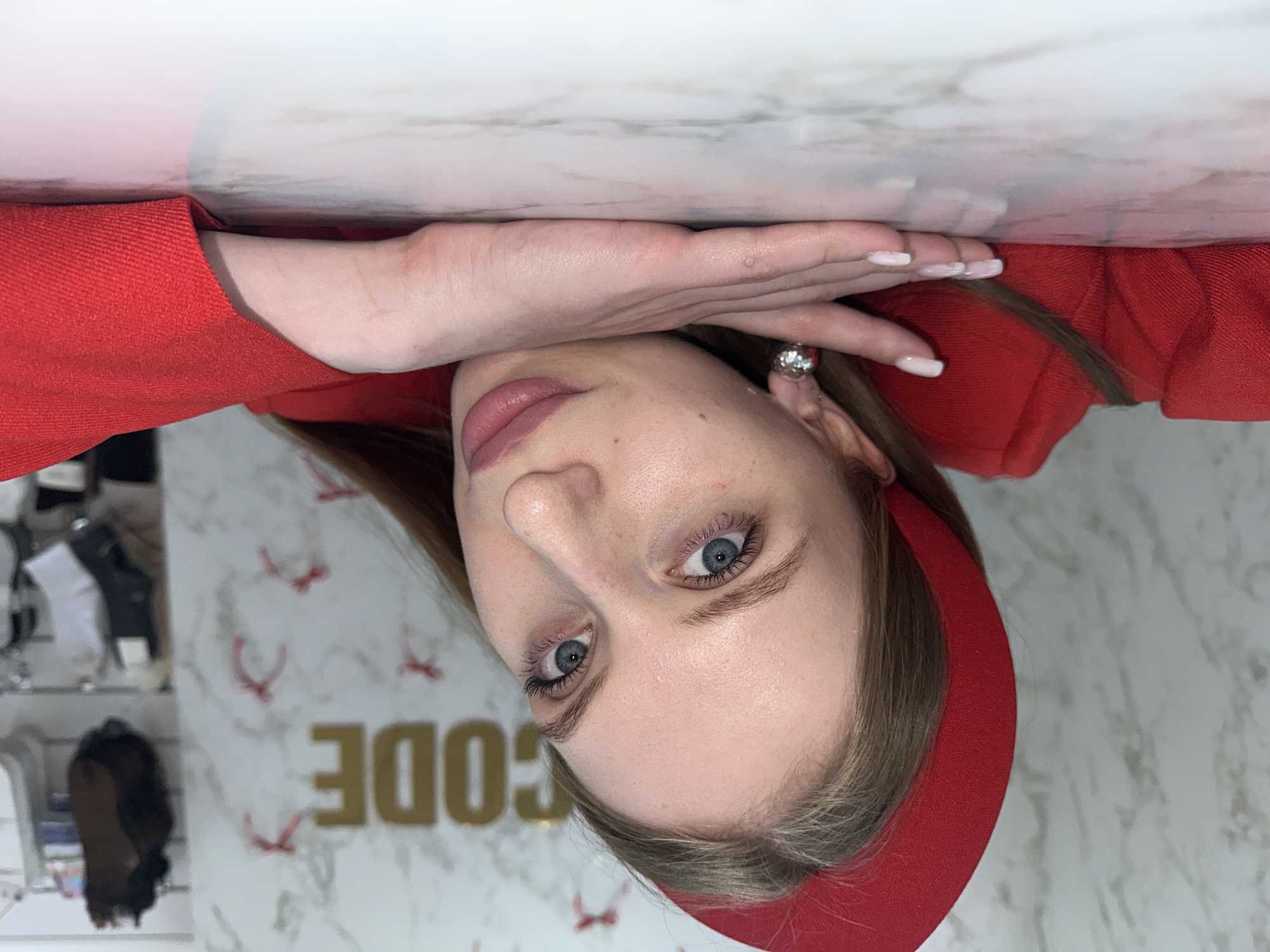A young woman with blue eyes and light brown hair is pictured in a close-up, slightly tilted portrait. She is wearing a red top and a matching headband. Her left hand is resting on a white marble-like surface. The background includes clothing items on shelves and gold lettering "CODE". The location is Chaikovsky, Russia.