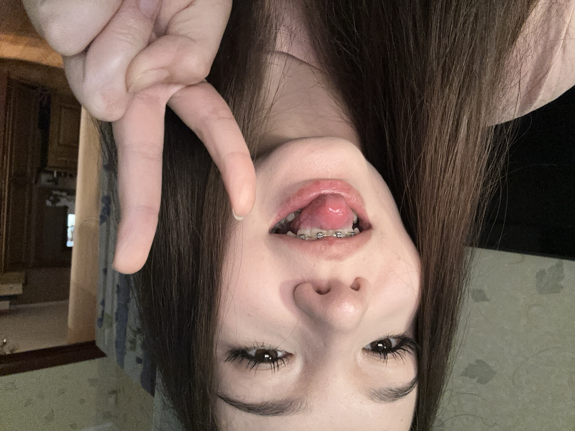 This close-up, inverted self-portrait captures a young person, likely female, from a low-angle perspective, possibly while they are lying down. The subject has long, straight dark brown hair that cascades towards the top of the frame. Their face is centered, featuring open brown eyes gazing directly at the camera, a visible nose, and an open mouth that reveals dental braces on the upper teeth and the tongue slightly extended. One hand is prominently displayed in the upper left quadrant of the image, making a "peace" or "V" sign.
The overall mood appears playful and casual, reinforced by the inverted pose and tongue gesture. The background indicates an indoor setting, specifically a room in what appears to be a home in Naberezhnye Chelny, Russia. In the upper left, hints of wooden cabinetry or furniture are visible, suggesting a kitchen or living area. To the left of the subject's face, a soft-toned, possibly patterned, light blue textile or wallpaper can be discerned. The lighting seems to be artificial and diffused, typical of an indoor environment, with no specific time of day clearly indicated. There is no visible text in the image.