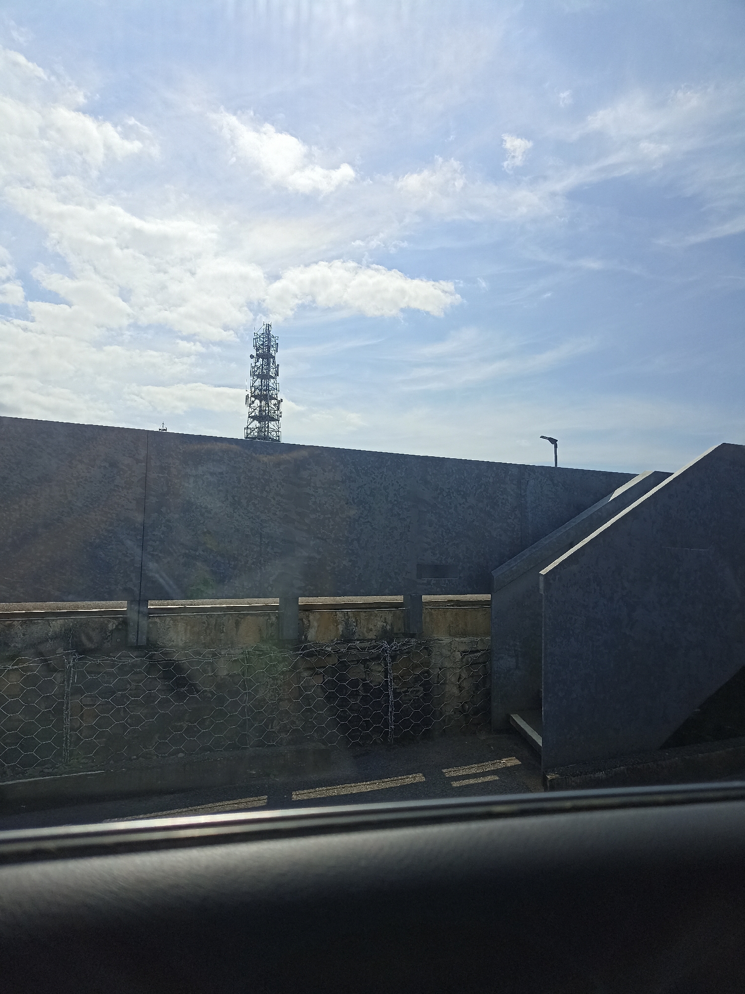 A cell tower stands tall against a bright, cloudy sky. In the foreground, a dark, textured surface, possibly a car interior, occupies the bottom portion of the frame. Beyond this, a grey, textured wall stretches horizontally, with a concrete structure featuring an angled upper section to the right. Below the wall, a wire mesh fence is visible, with a glimpse of a paved area and some greenery beyond. The scene appears to be an outdoor urban or industrial setting.