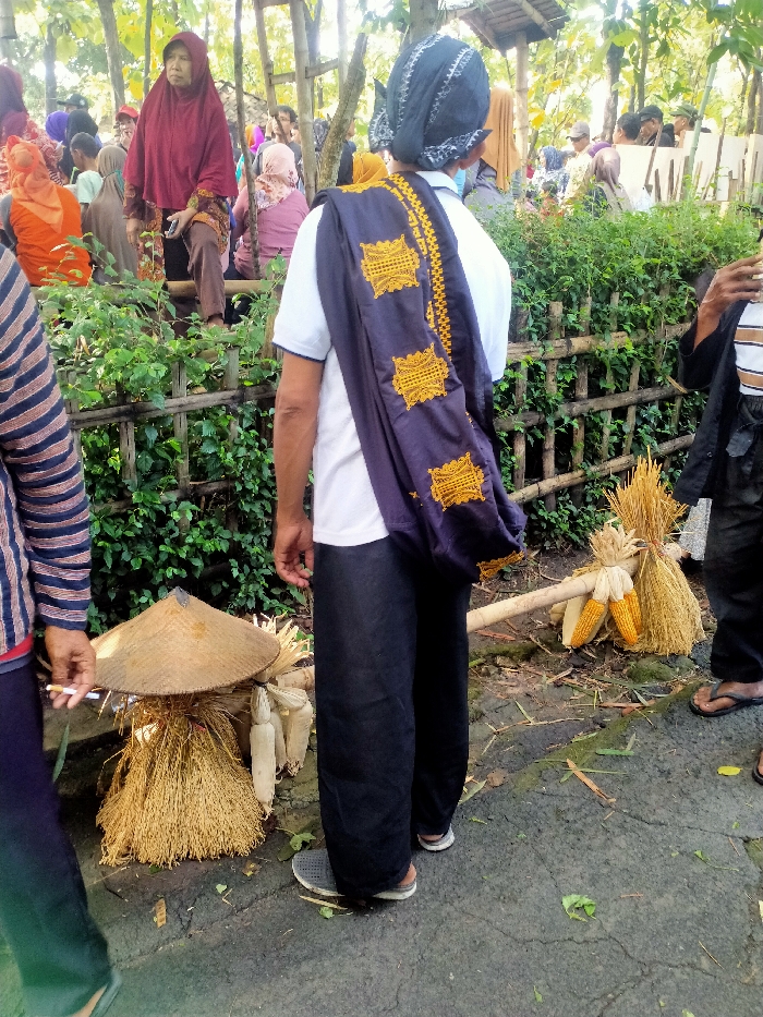 The scene is in Masaran, Indonesia, at the coordinates -7.472 latitude, 110.956 longitude. It is a street scene with a group of people gathered near a bamboo fence and a thicket of trees. A man wearing a white shirt and black pants with a blue and gold patterned shawl stands on the right, facing the fence. He is looking toward the left of the frame. He is standing near a bamboo stick that has a bundle of dried corn stalks attached to it. To the left of the man is a large straw hat placed on the ground with a bundle of dry stalks beneath it. In the background, there are a multitude of people, some of whom are wearing traditional clothing. They are all standing or sitting and appear to be socializing or shopping. There is also a bamboo fence that serves as a border for the scene. The ground is paved with gray asphalt and there are various scattered leaves and bits of debris.