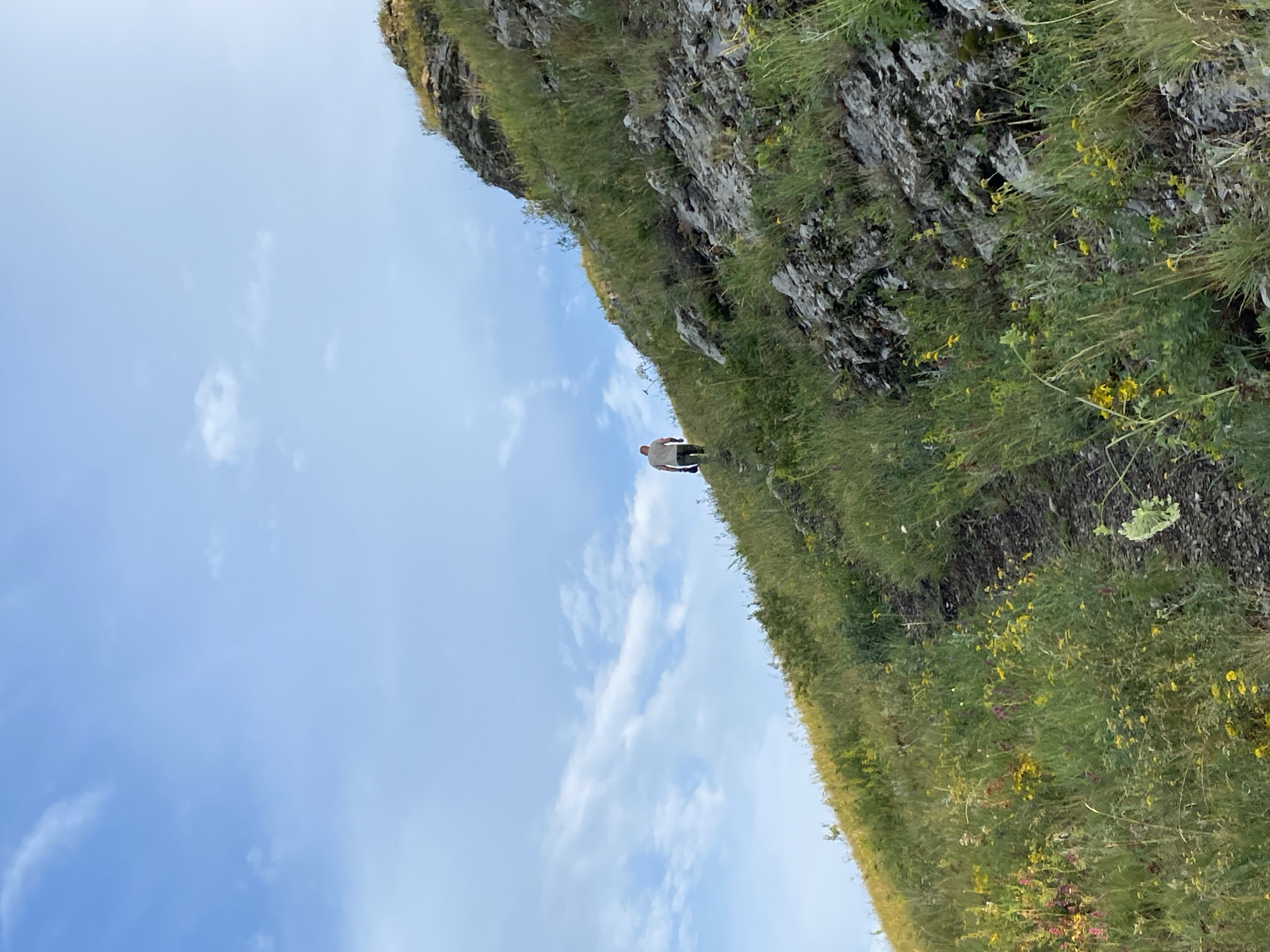 The image depicts a person walking up a steep, grassy hillside on a clear day. The sky is a bright blue with a few wispy white clouds. The hillside is covered in green grass and various wildflowers in yellow and pink hues. Rocky outcrops are visible along the slope. The person, wearing a light-colored t-shirt and dark pants, is walking away from the camera, ascending the path. The scene appears to be an outdoor natural landscape.