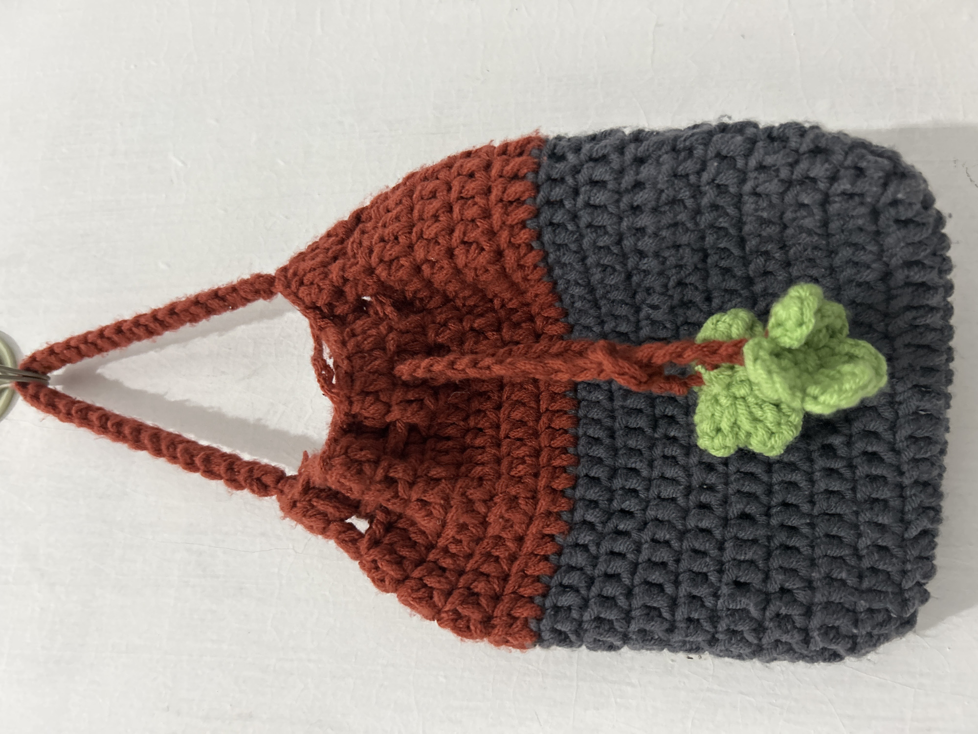 A handcrafted crocheted pouch lies against a plain, light-colored background. The pouch features a two-tone design: the upper section is a rich rust orange, while the larger lower section is dark gray. A crocheted drawstring, matching the rust orange color, is threaded through the top of the pouch. One end of the drawstring is attached to a silver-colored metal ring, while the other end is tied, securing a small, light green crocheted clover or leaf appliqué against the dark gray portion of the pouch. The crochet stitches are clearly visible, showcasing the texture of the yarn. The item is stationary, likely positioned for photography, with no activity taking place. There is no visible text. The lighting is even, suggesting an indoor setting.